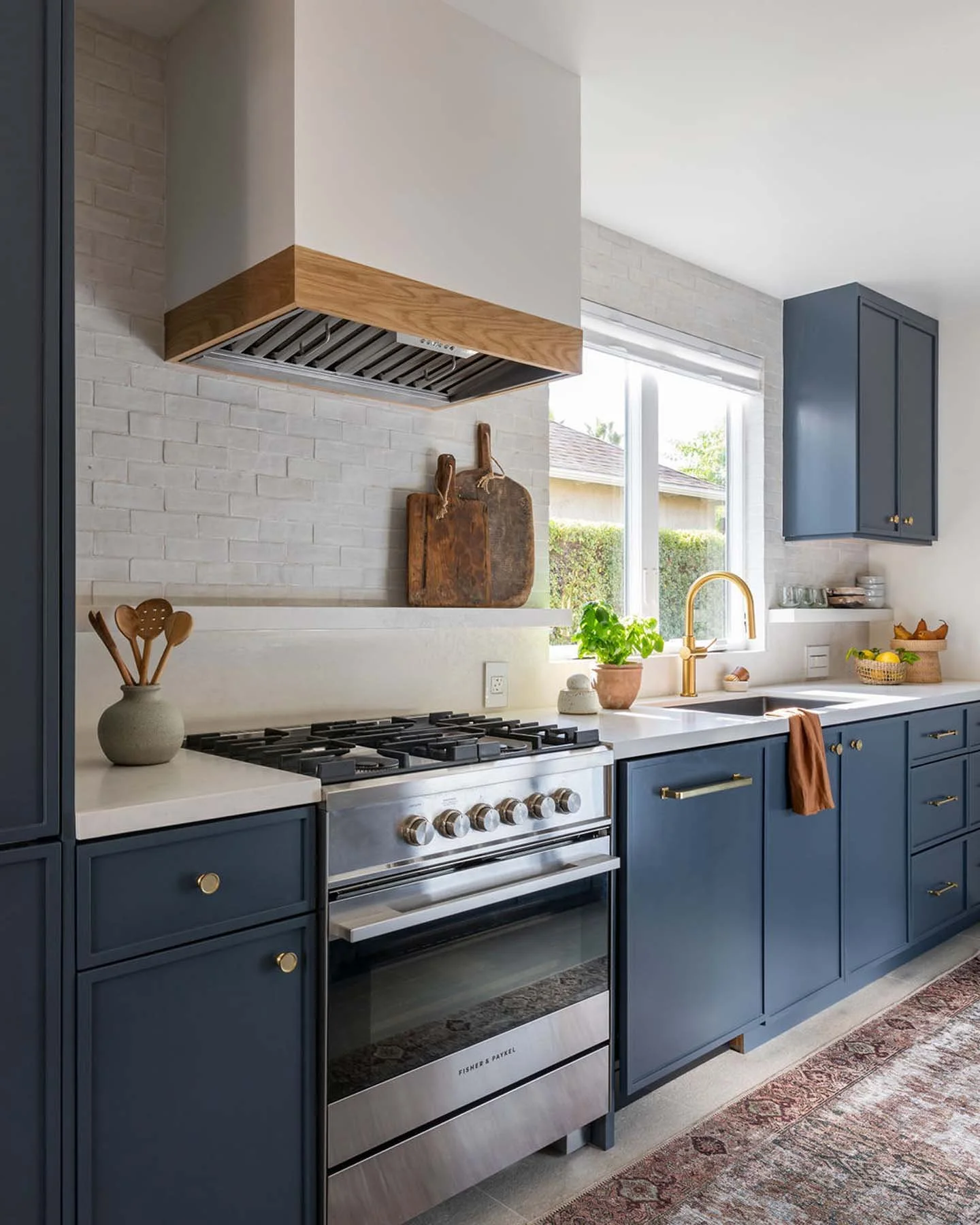 Modern kitchen with blue cabinets, stainless steel stove, white countertops, and decorative items including cutting boards, a potted plant, and bowls. There is a window with sunlight and a patterned rug on the floor.