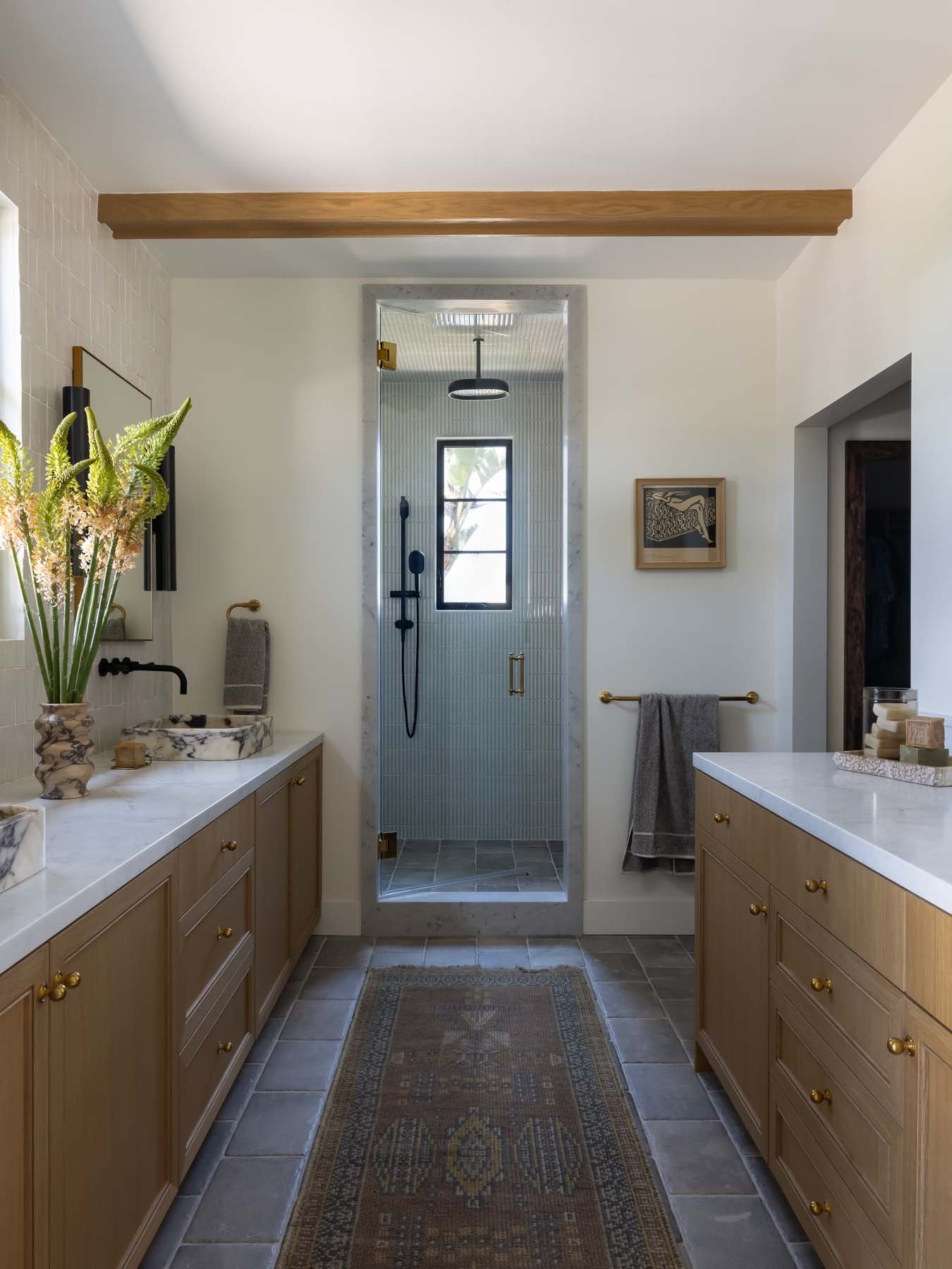 A bathroom with double vanities, wooden cabinets, marble countertops, decorative vases, a rug, and a walk-in shower enclosed with glass.