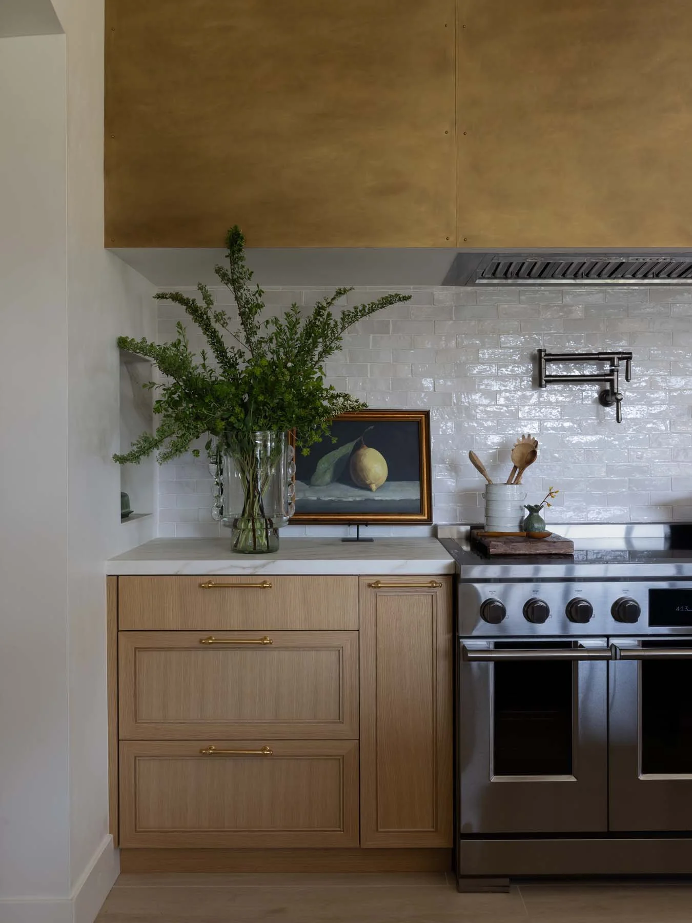 Kitchen with light wood cabinetry, a large flower arrangement in a glass vase, a still life painting, and a stainless steel stove against a white brick backsplash.