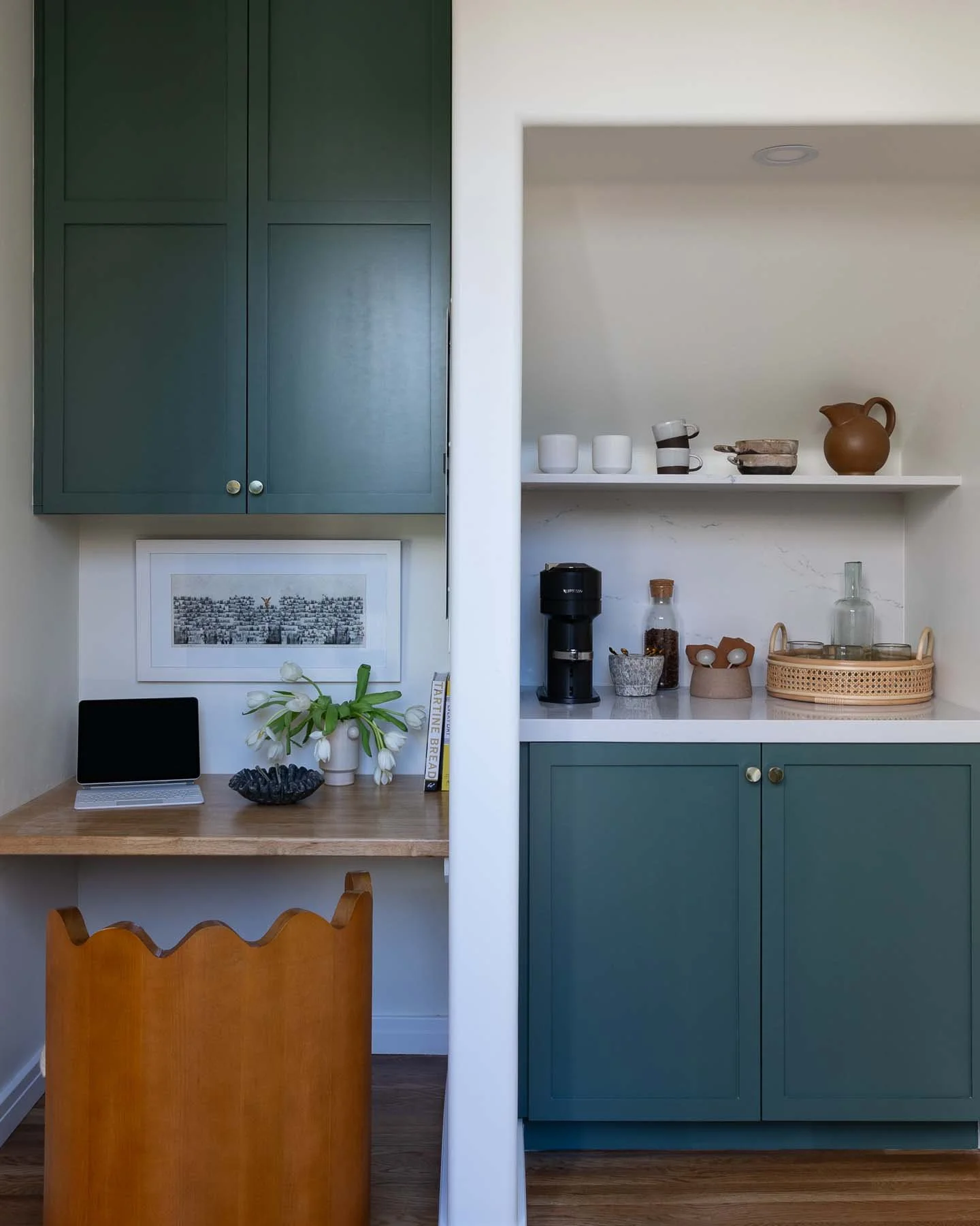 Split view of a kitchen area with green cabinetry, open shelving, and a workspace with a wooden desk, a tablet, flowers, and books on the left side, and shelves with cups, jars, and a pitcher on the right side.
