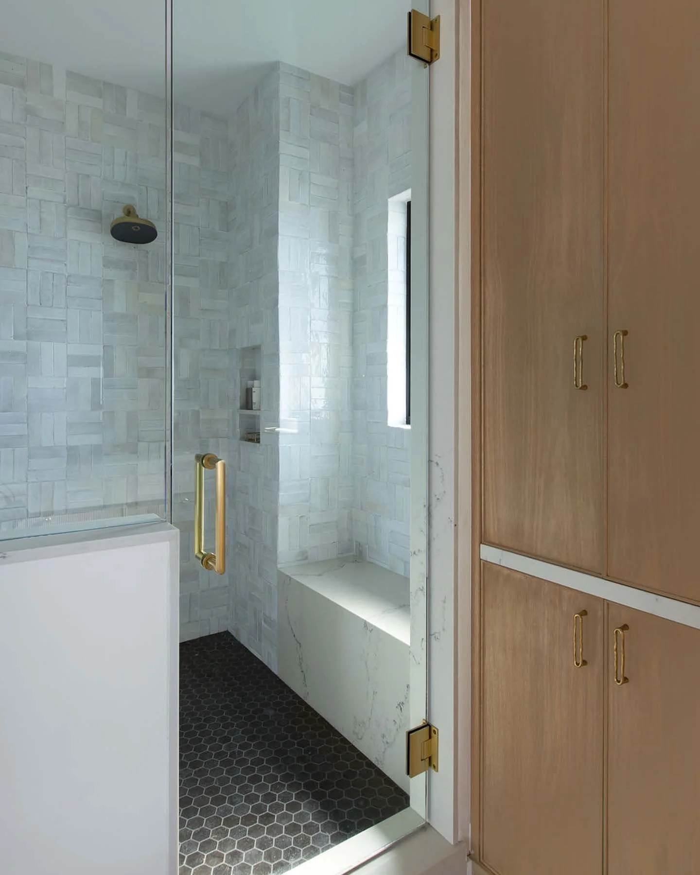 Indoor bathroom shower with frosted glass door, black hexagonal floor tiles, white marble walls, and a window with natural light. Wooden cabinetry with gold handles adjacent to the shower.