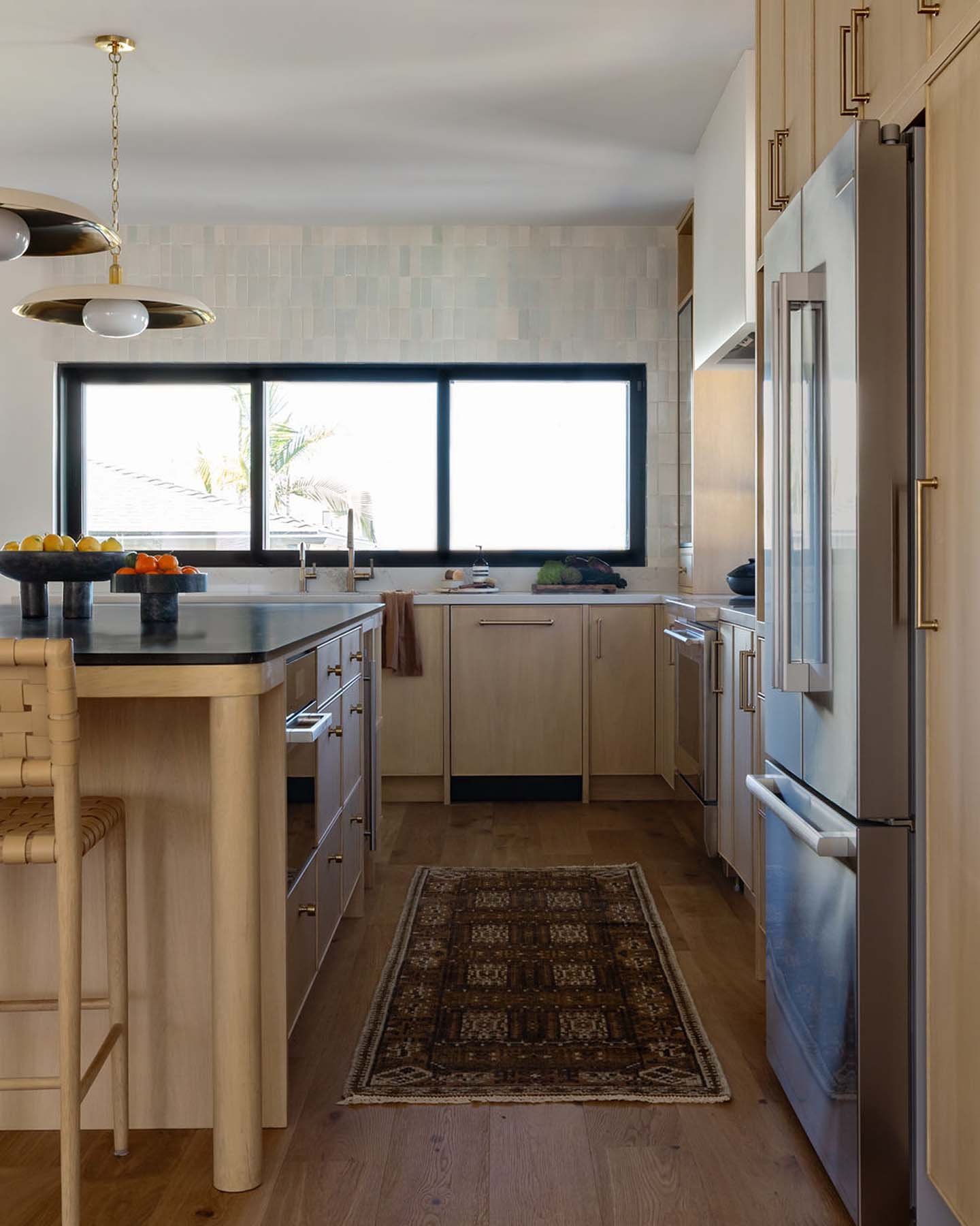 A cozy kitchen with wooden cabinets, a large window above the sink, and a patterned rug on the wooden floor. There is a kitchen island with fruit bowls and a light fixture hanging from the ceiling.