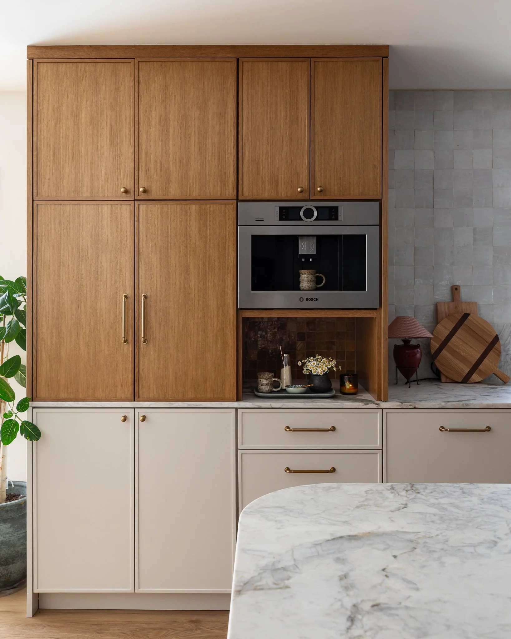 Modern kitchen with wooden upper cabinets and white lower cabinets, marble countertop, built-in microwave, and decorative items on open shelf, with a section of gray tiled wall and part of a marble kitchen island in the foreground.