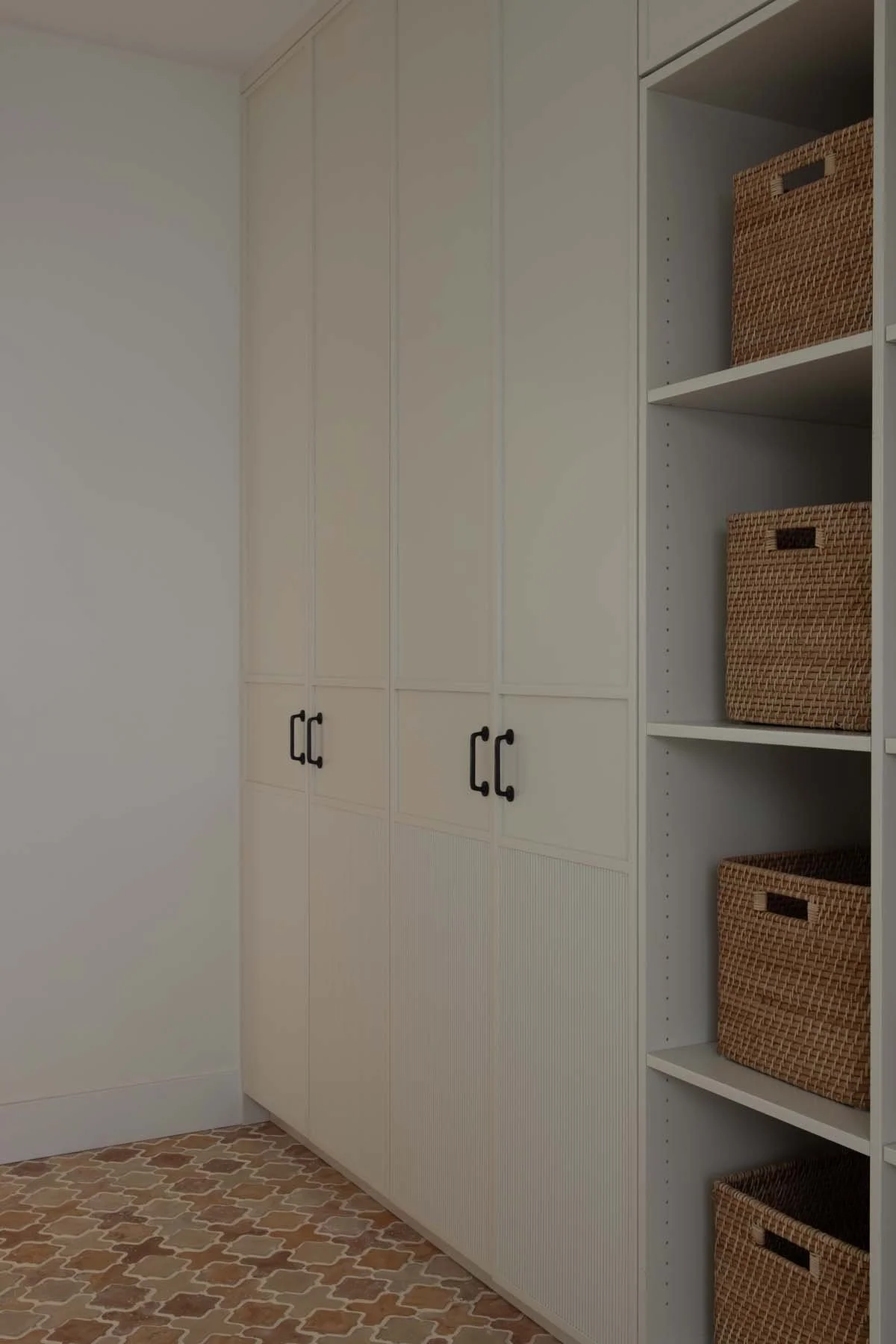 A closet with white cabinets and black handles, alongside white shelves holding woven baskets, a corner of a patterned tile floor, and a plain white wall.