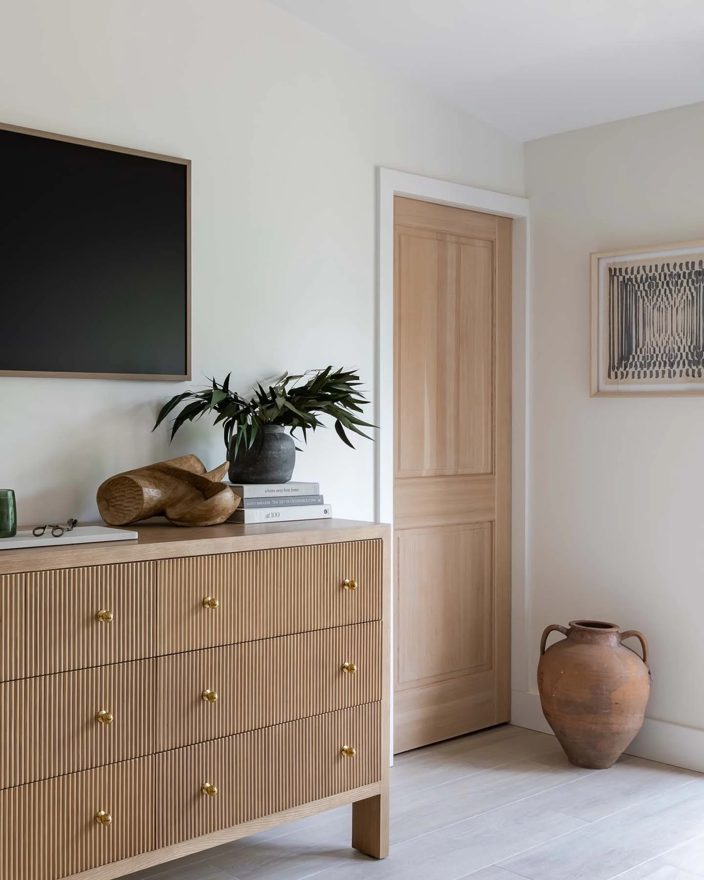 A wooden sideboard with vertical grooves and gold knobs in a bright room. On top, there is a dark vase with green leaves, a carved wooden object, and a stack of books. To the right, there's a large terracotta vase on the floor. The wall has a black frame with art and a wooden door.