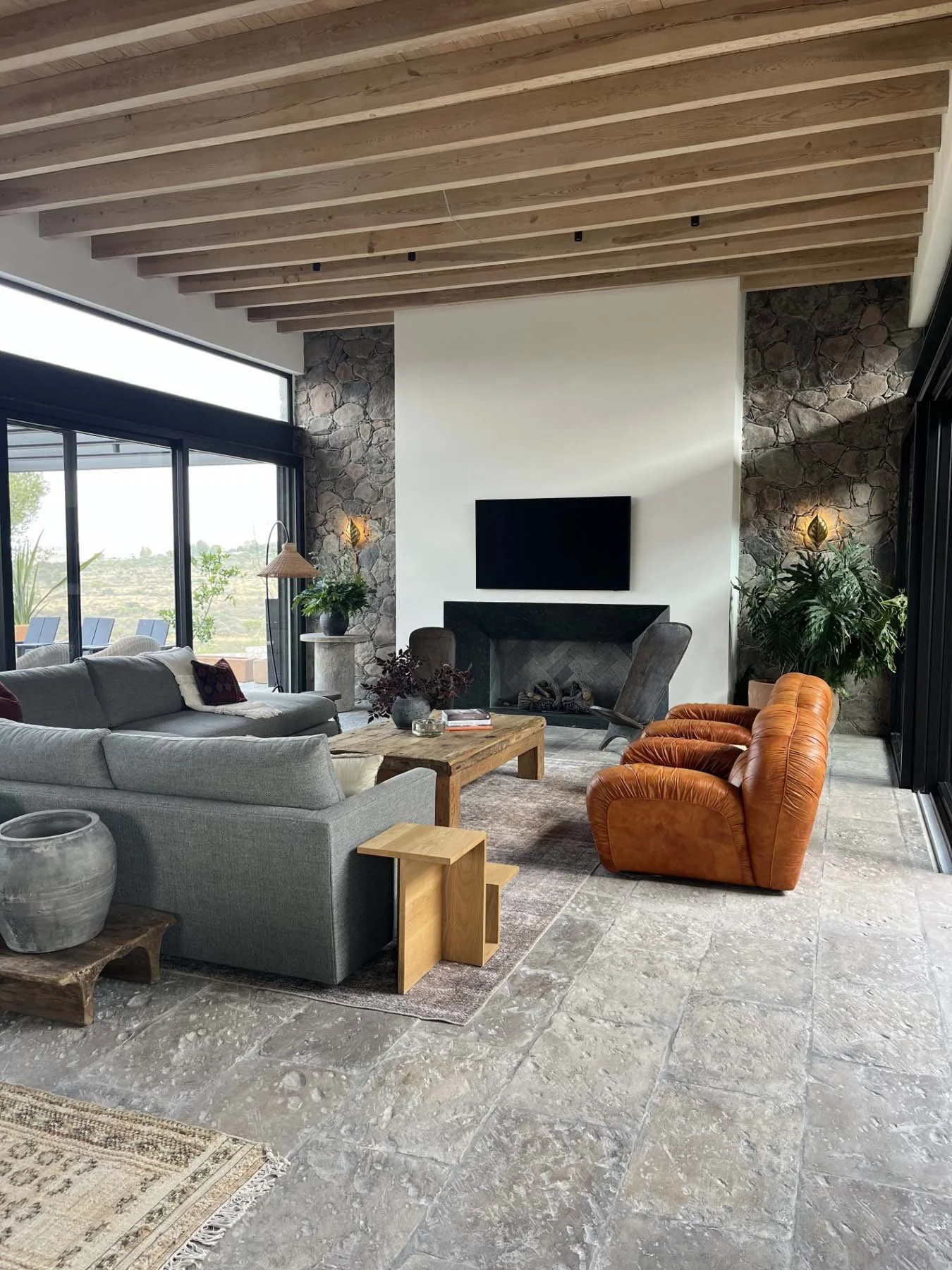 Living room with wood-beamed ceiling, stone accents on walls, large windows, and modern furniture including a grey sofa, orange armchairs, a wooden coffee table, and plants.