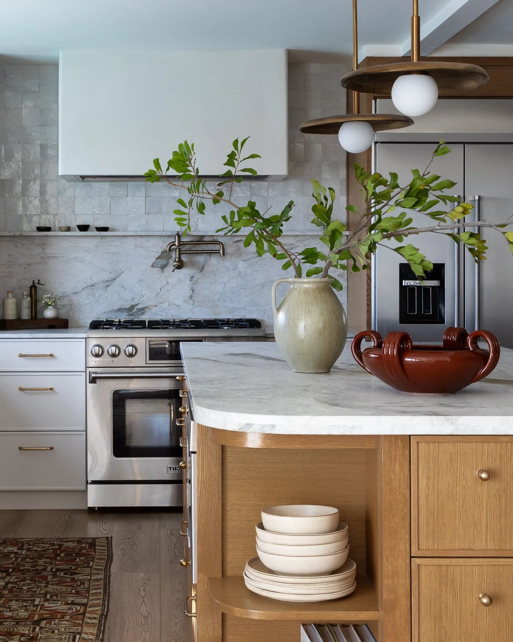 A kitchen with a marble island countertop, a decorative bowl, shelves with plants, and a stainless steel refrigerator.