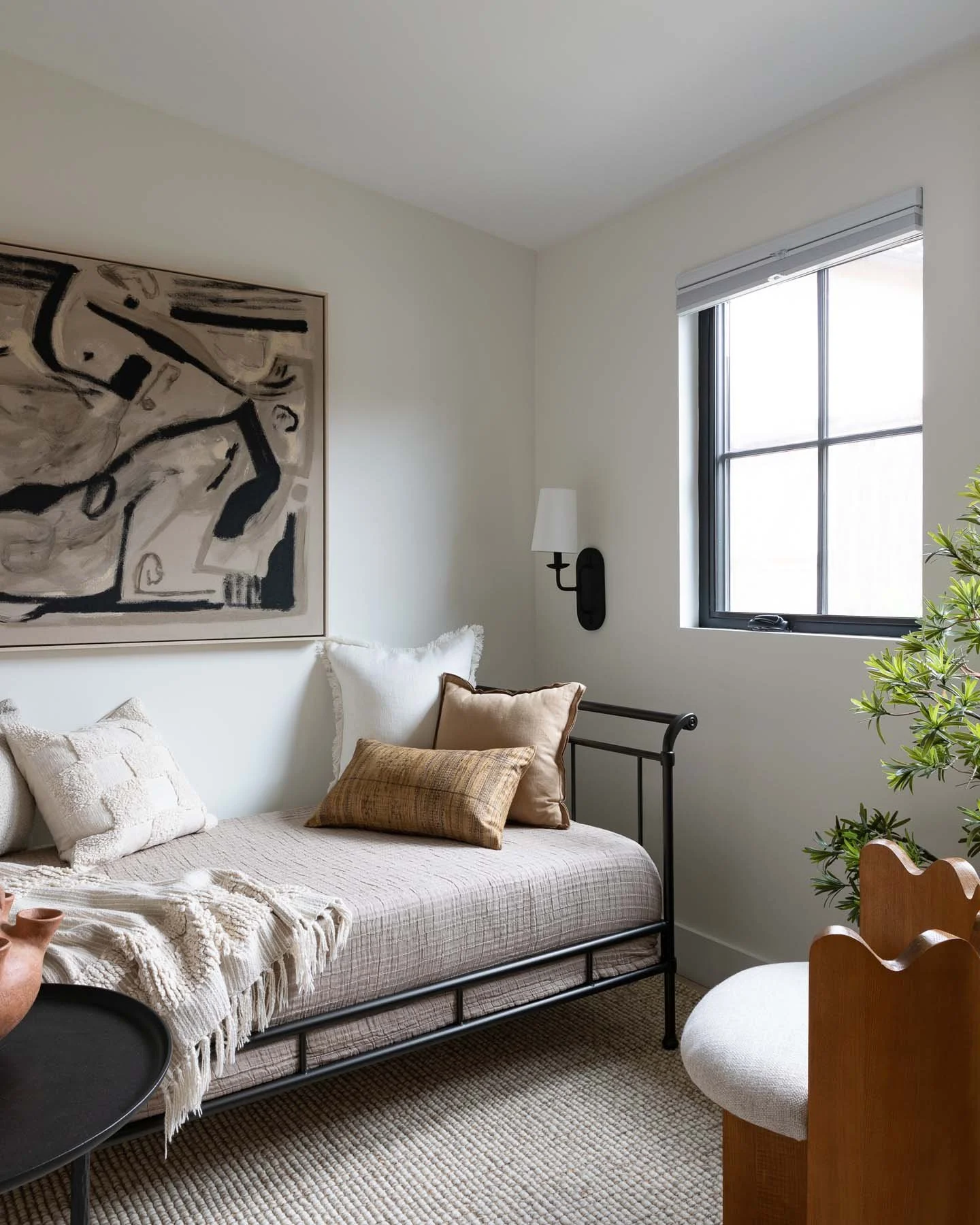 A cozy bedroom corner with a minimalist bed dressed in a neutral bedspread, decorated with assorted pillows. There is a doorway or window with a black frame, and a contemporary black wall sconce with a white shade. A large abstract black and white painting hangs above the bed, and a leafy green plant is partially visible on the right side. The room features a beige rug on light-colored wood flooring and a small black round tray table.