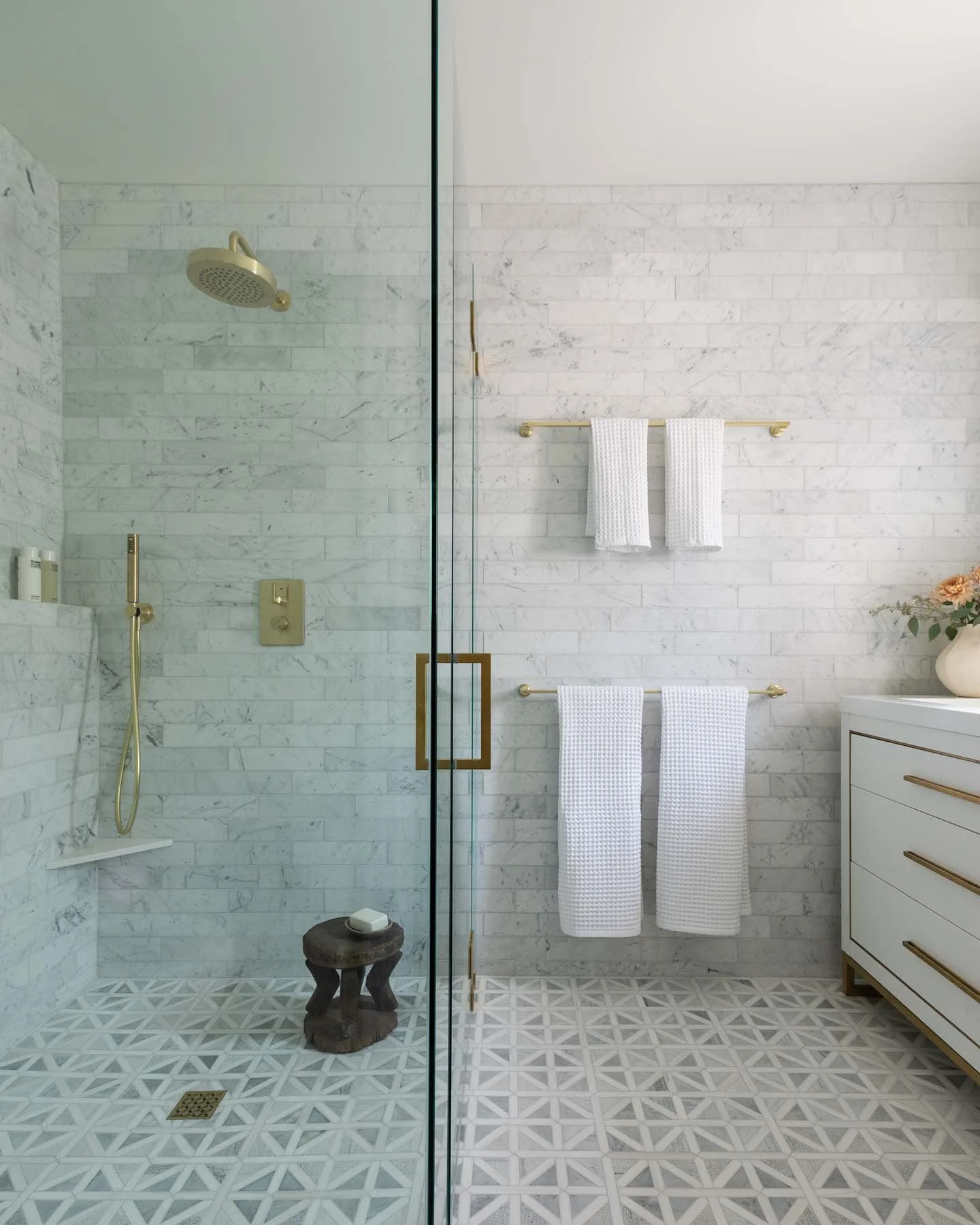 Modern bathroom with a walk-in shower featuring white marble tiles, a gold showerhead, and a gold handheld shower. There are white towels hanging on a gold towel rack, a white dresser with gold handles, a small wooden stool, and a flower vase with pink flowers.