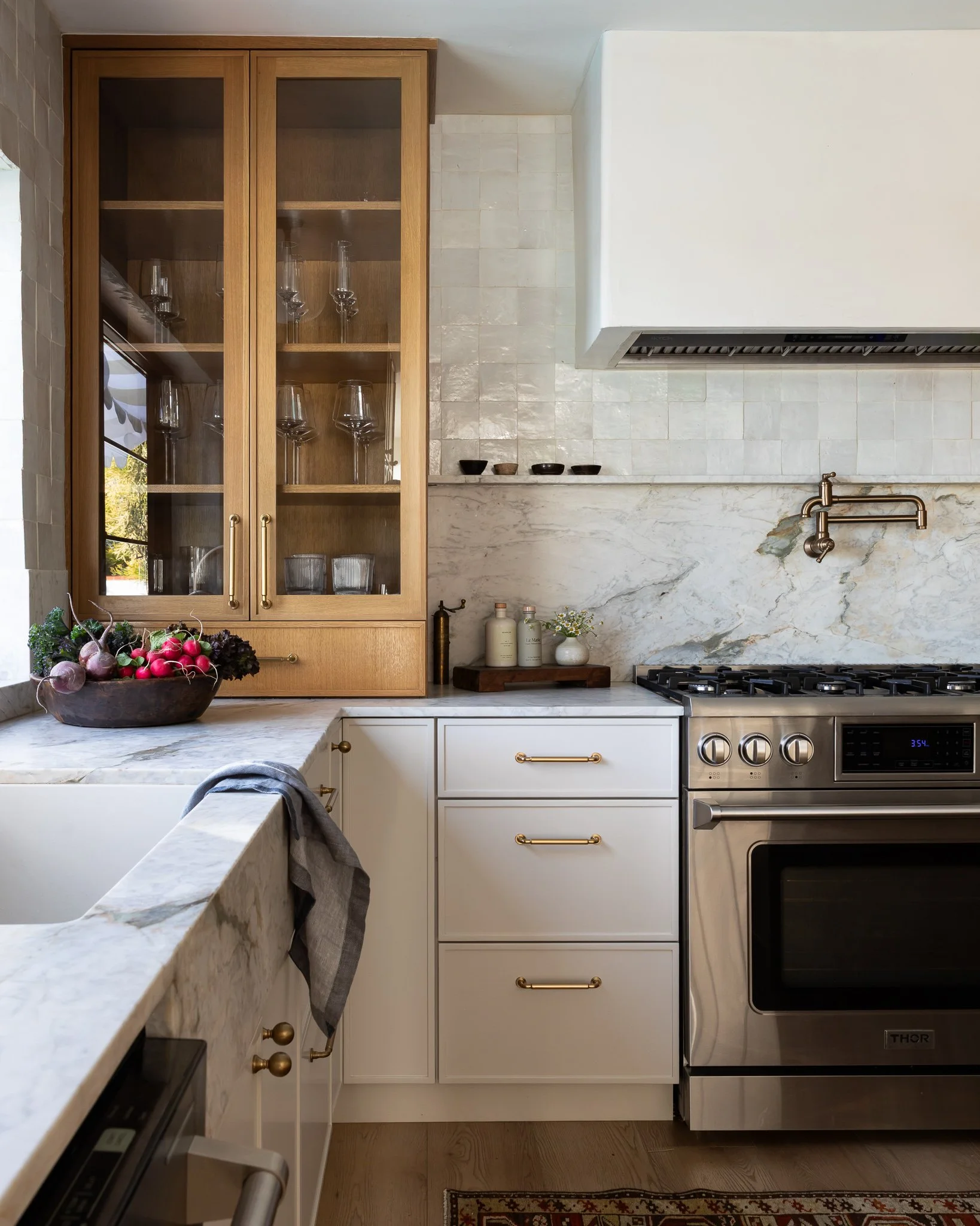 Modern kitchen with white cabinets, marble countertops, a stainless steel oven, and a custom marble backsplash. An open wooden cabinet displays glassware, and a bowl of radishes is on the counter.