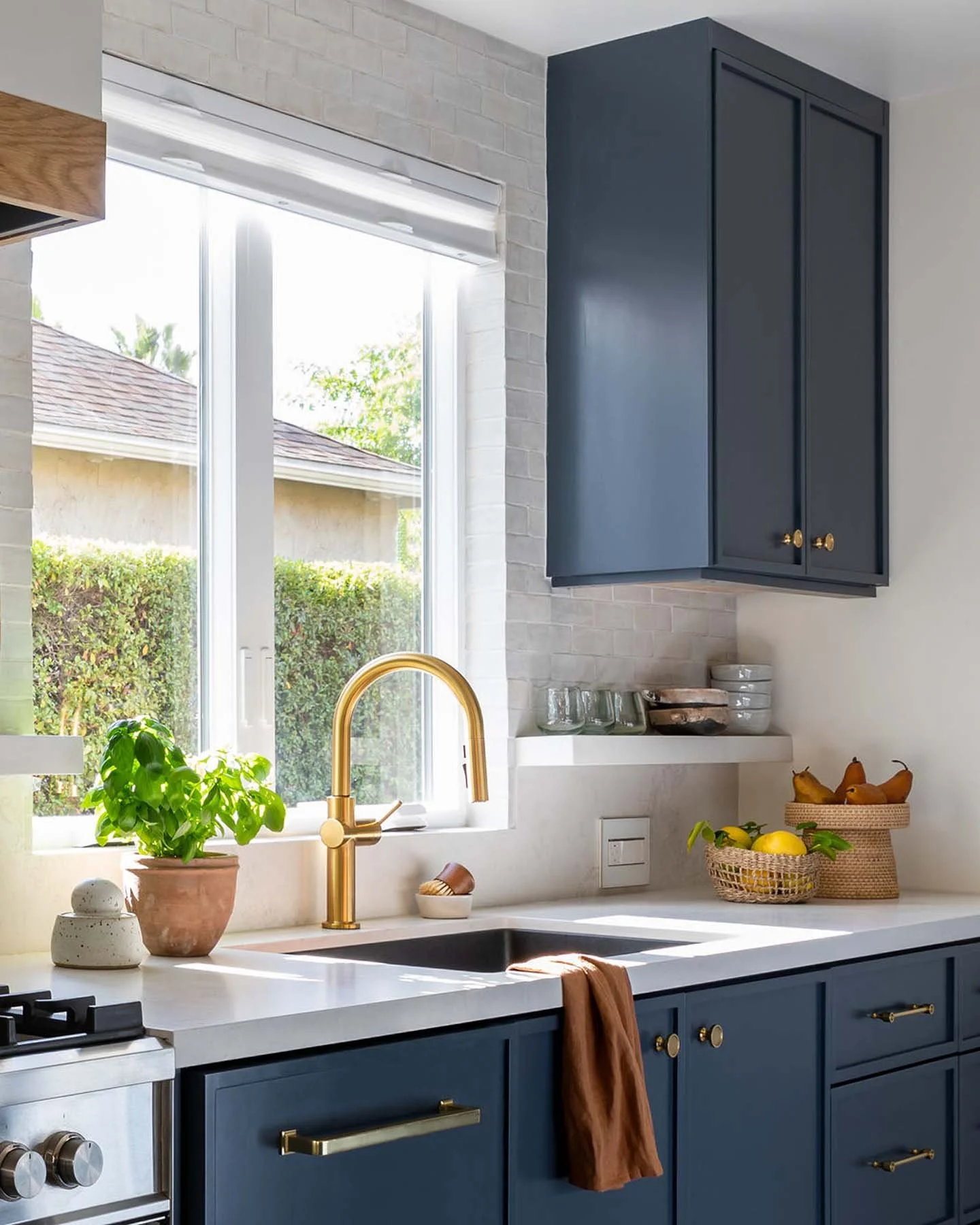 Kitchen with blue cabinets, white countertop, gold faucet, window with view of outside greenery, potted basil, decorative stones, and baskets of fruits on the counter.