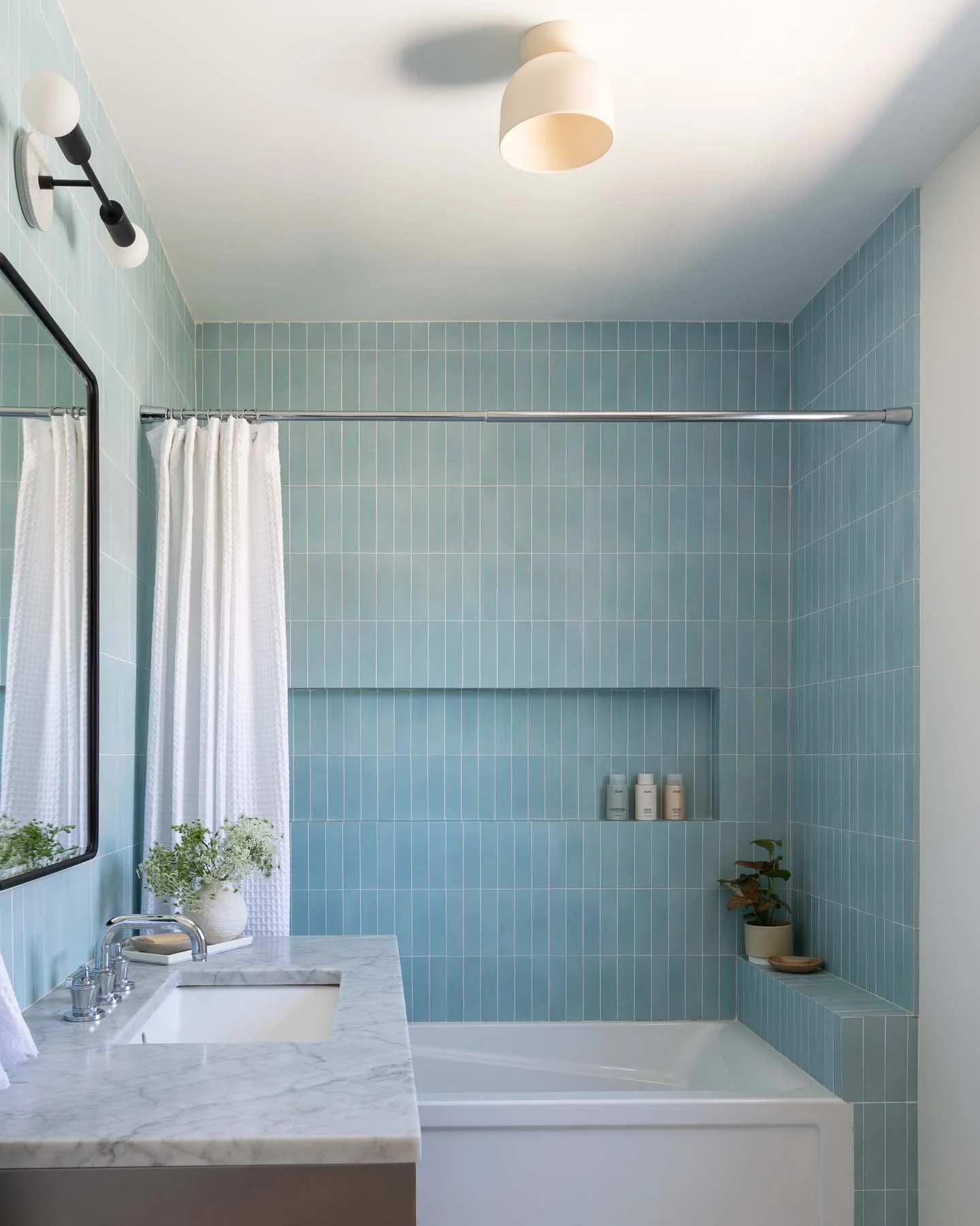 A modern bathroom with light blue tiled walls, a mirror, a white marble countertop with a sink, a potted plant, and a shower area with a curtain and built-in shelves holding bottles.