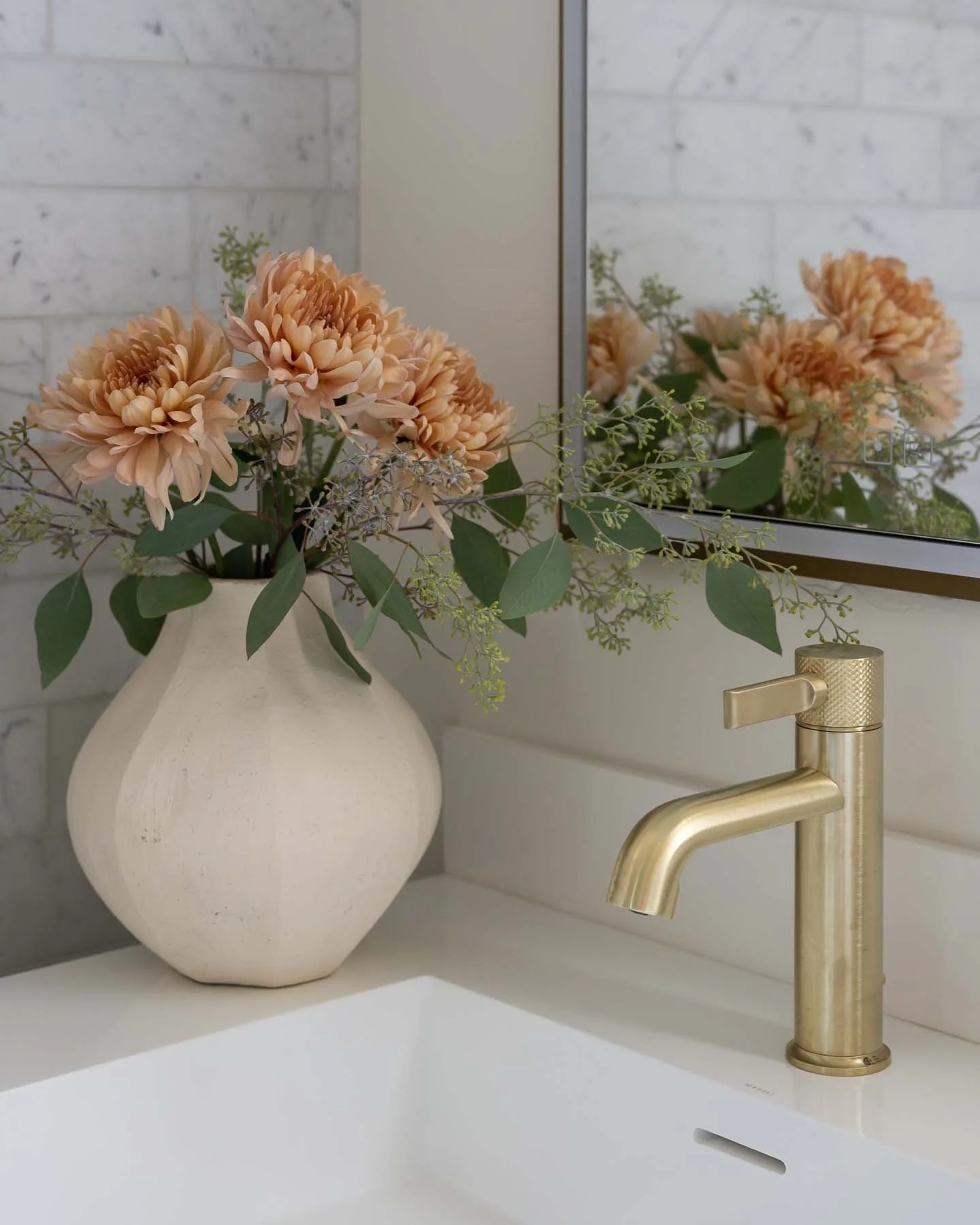 A white vase with pink and peach flowers and green leaves on a bathroom sink. A gold faucet is visible next to the vase, and a mirror reflects part of the flowers and the white brick wall.