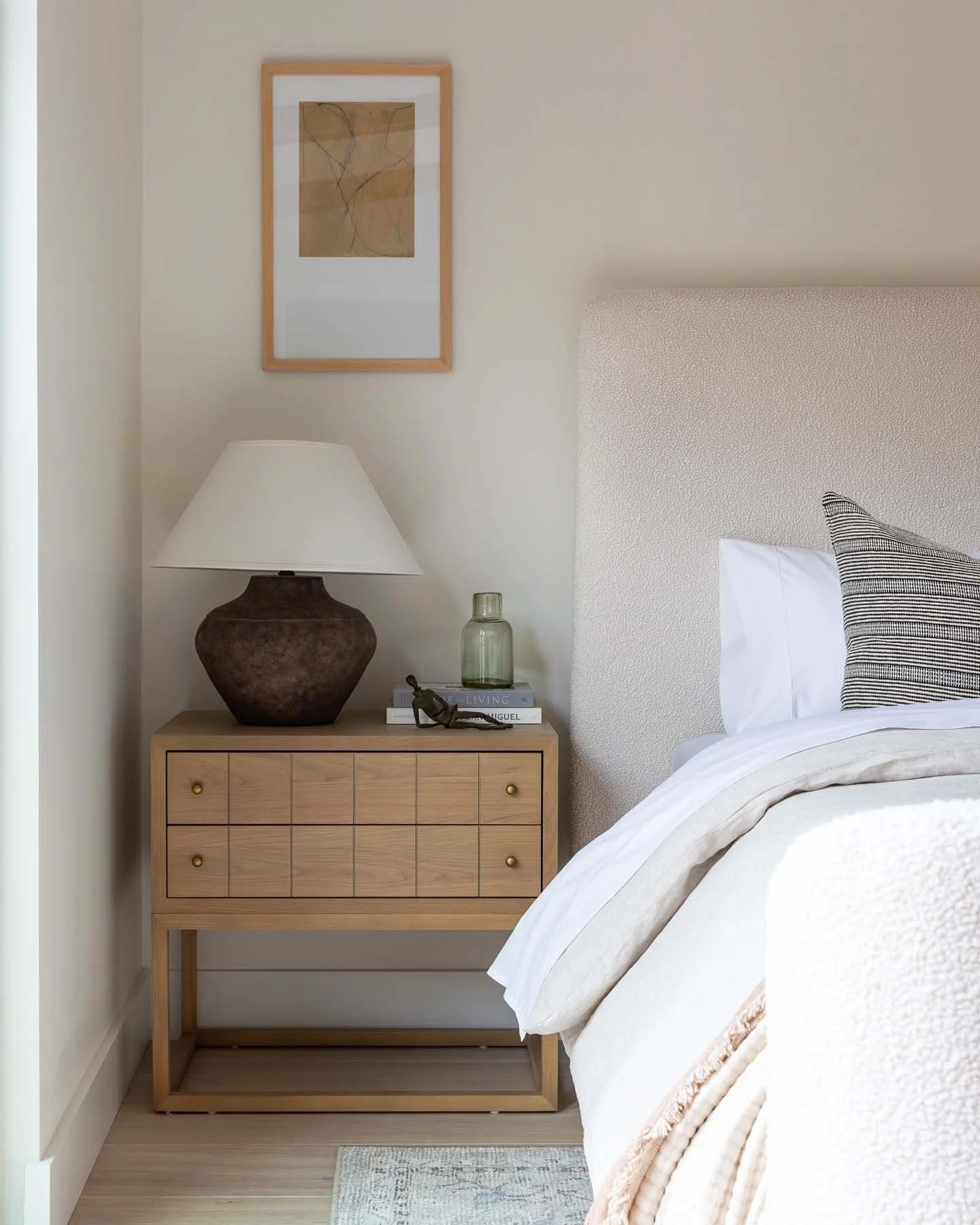 A bedroom corner with a bed, a wooden nightstand, a table lamp, a green glass bottle, a stack of books, and a small decorative object. There is a framed abstract art piece on the wall above the nightstand.