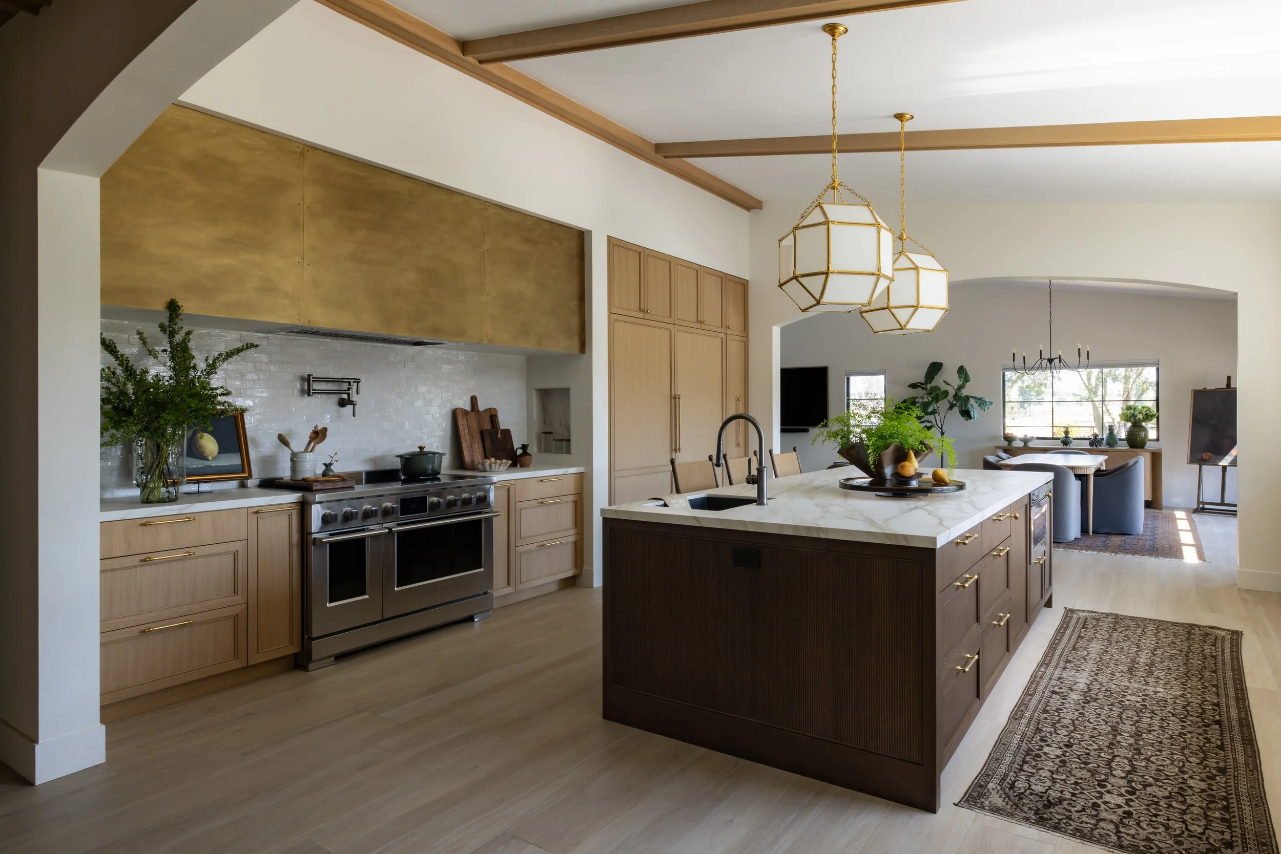 Modern kitchen with wooden cabinets, stainless steel oven, marble island with a sink, hanging pendant lights, and visible dining area with large windows and a chandelier.