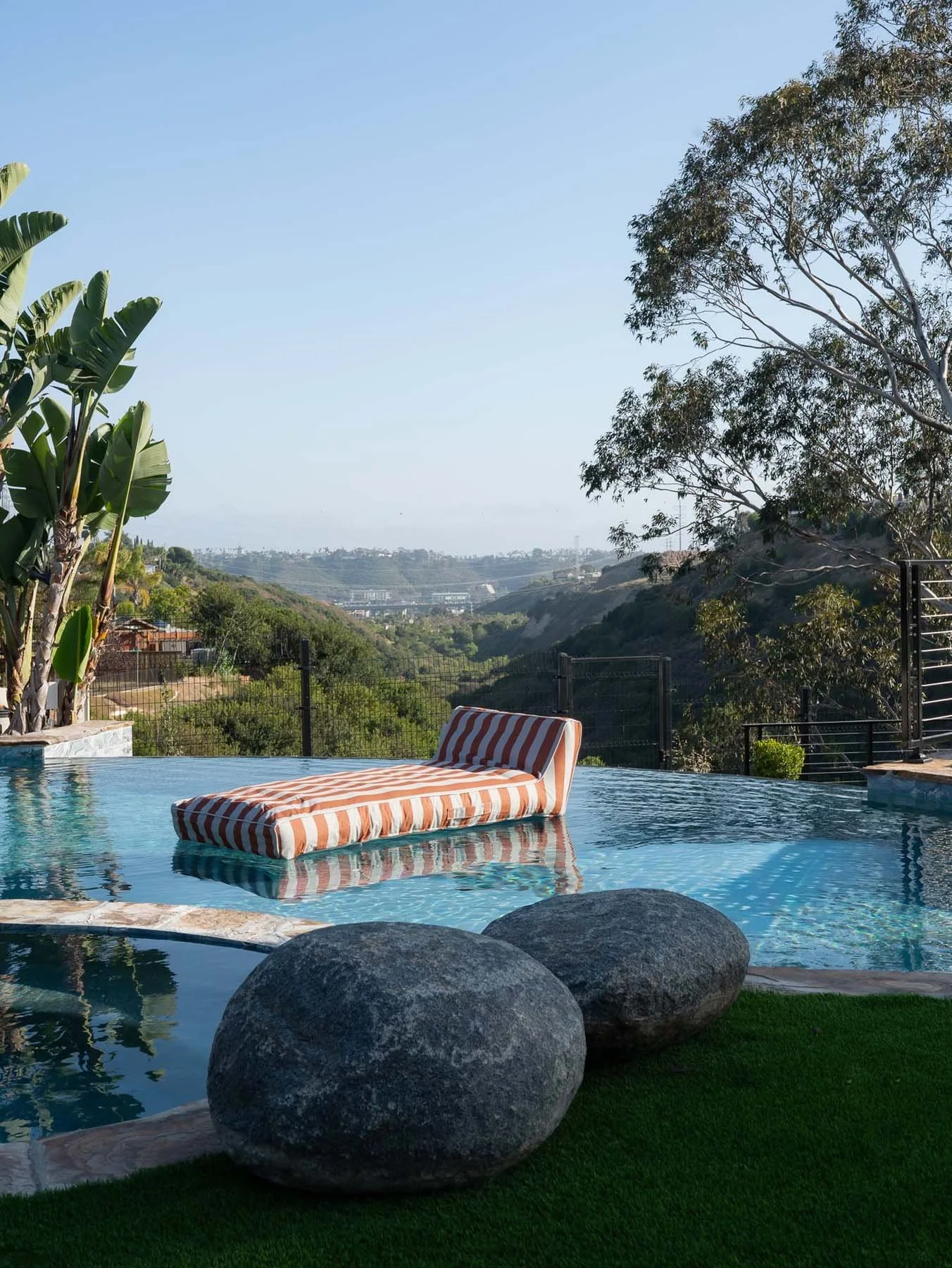 A swimming pool with a striped lounge chair floating on the water, surrounded by large rocks and lush greenery, overlooking hills and a distant cityscape under a clear blue sky.