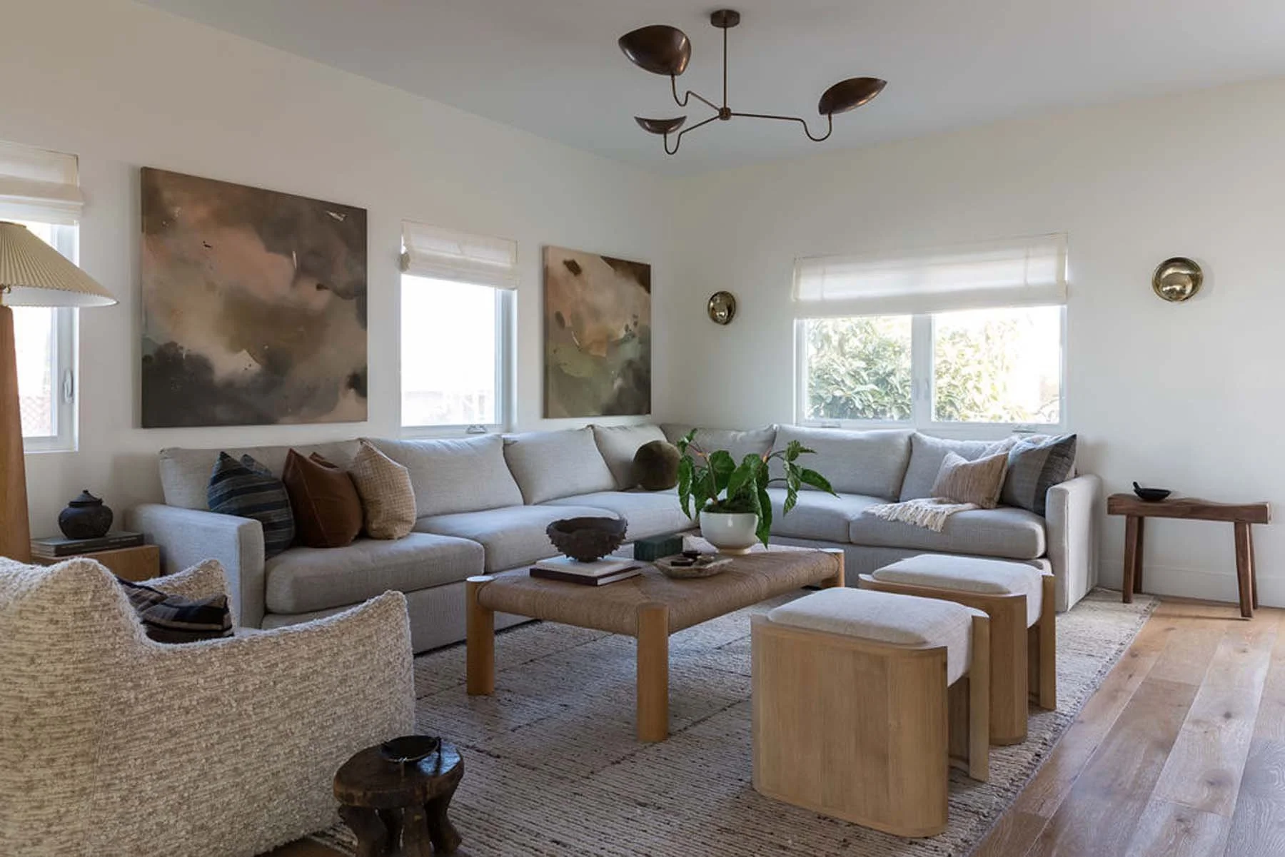 Living room with beige sectional sofa, wooden coffee table, abstract wall art, window blinds, indoor plants, and wooden side tables, with light-colored walls and hardwood flooring.