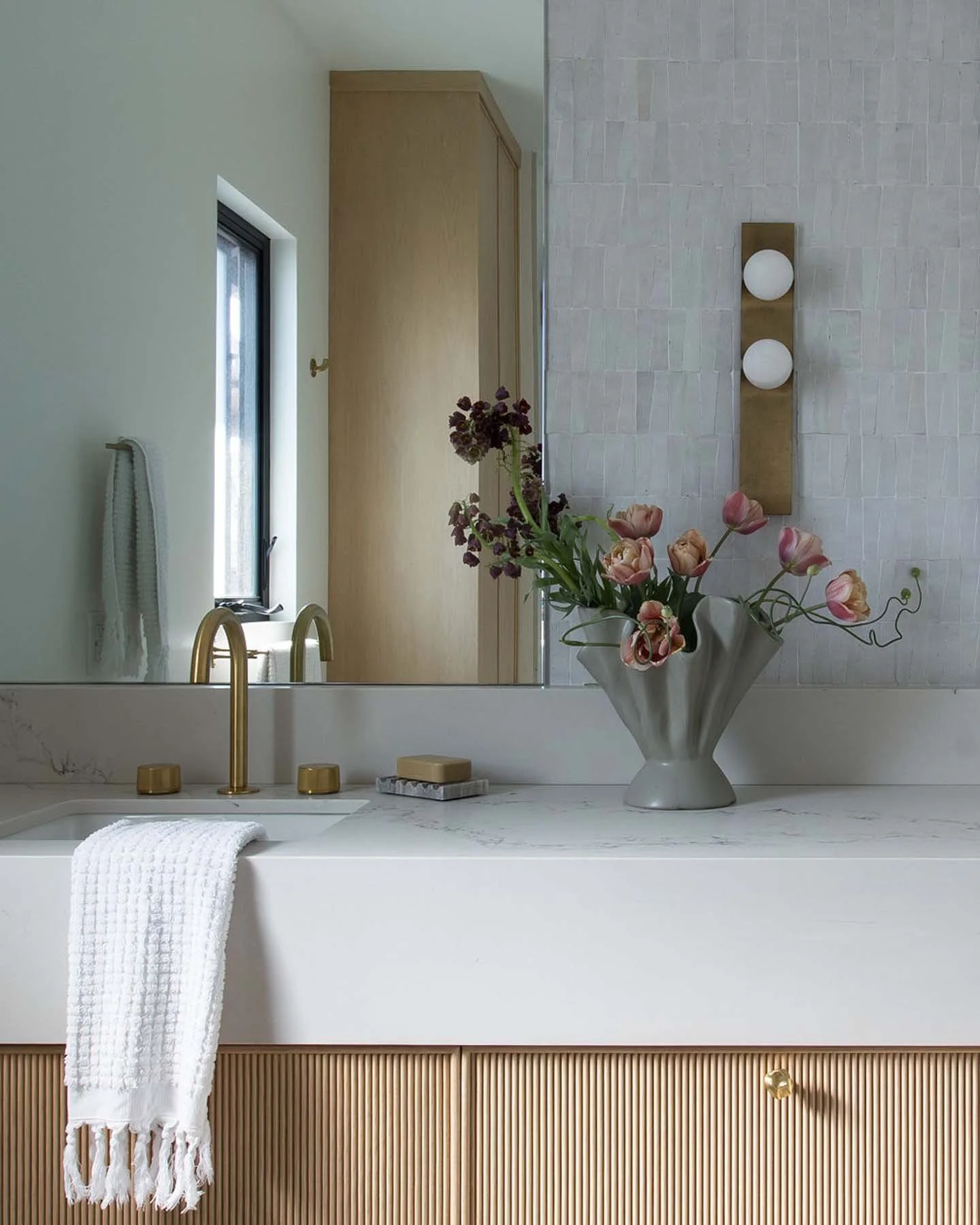 A bathroom vanity with a marble countertop, gold faucet, and a white towel hanging off the front. Behind the counter, there is a large mirror, a vase of pink flowers, and a light fixture with two round white bulbs. A window with a black frame is reflected in the mirror.