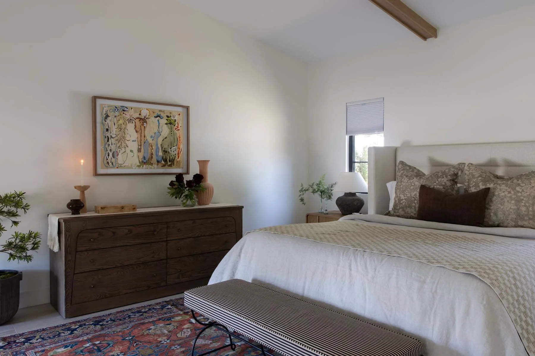A bedroom with a white bed, patterned pillows, a wooden dresser with decorative items, a framed artwork, a window with blinds, and a bench at the foot of the bed.