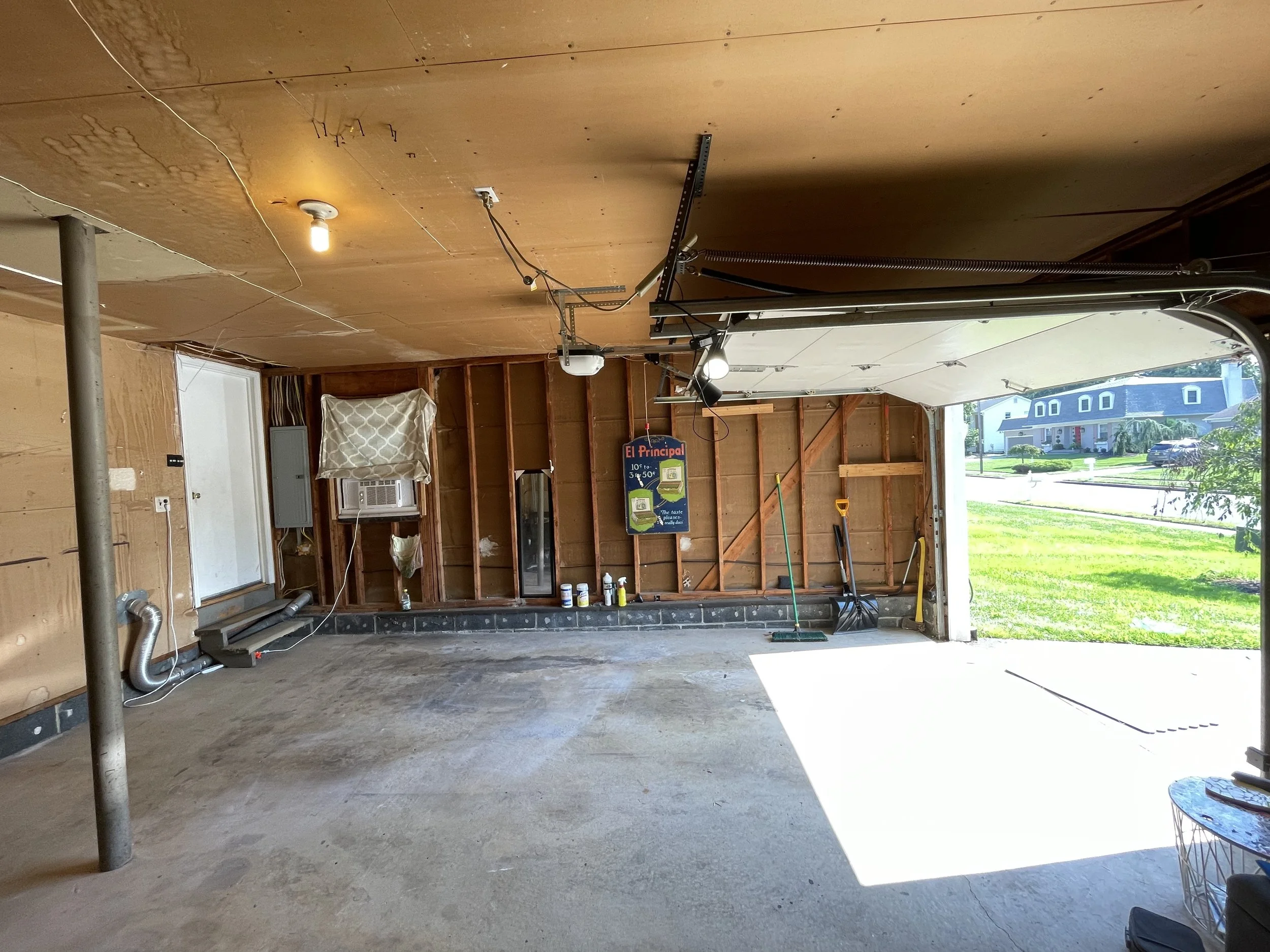 View of an unfinished garage or workshop with exposed wooden walls, concrete floor, and garage door open to outdoors. Some tools, equipment, and supplies are visible, including a small sign and an air conditioning unit.