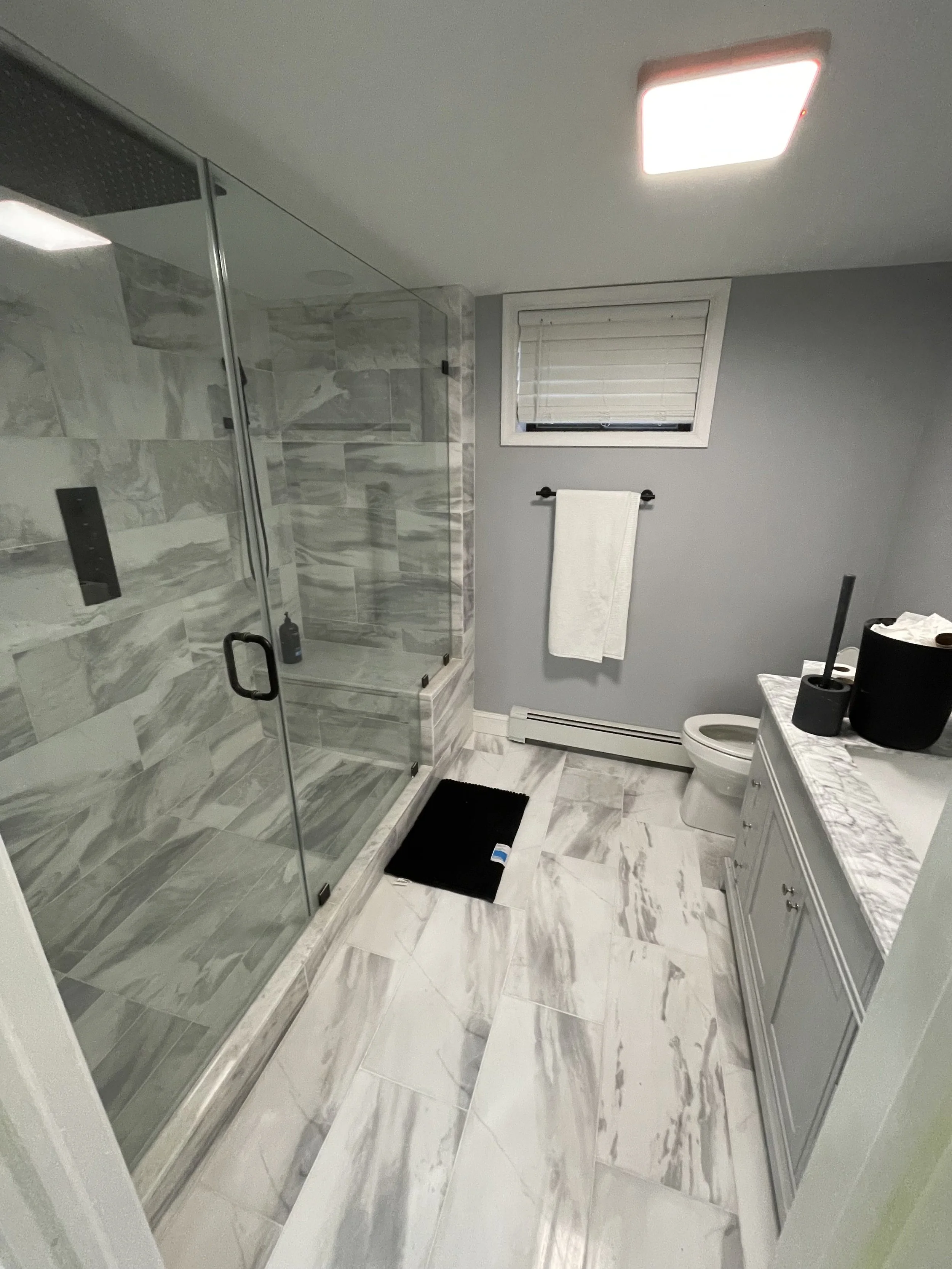 Modern bathroom with a glass-enclosed shower with gray marble tiles, a white vanity with a marble countertop, a window with white blinds, and a towel hanging on a black towel bar.