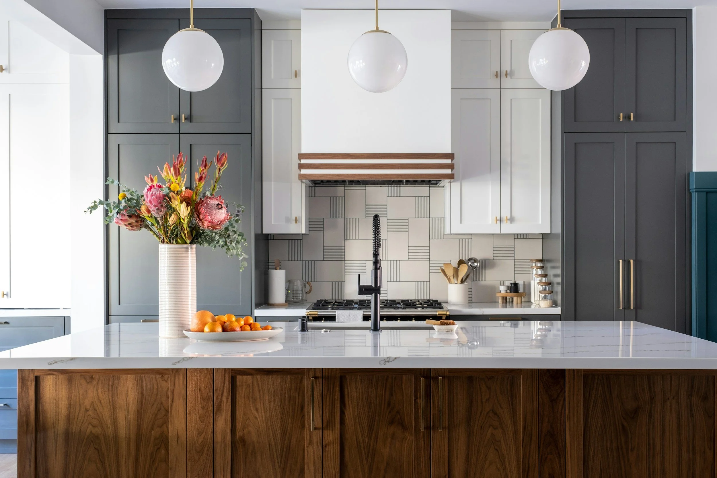 Modern kitchen with dark gray cabinets, white marble island, and a white tile backsplash. There are three globe pendant lights hanging over the island. A vase with colorful flowers and a bowl of oranges are on the island. There are wooden accents on the island and gold hardware on the cabinets.