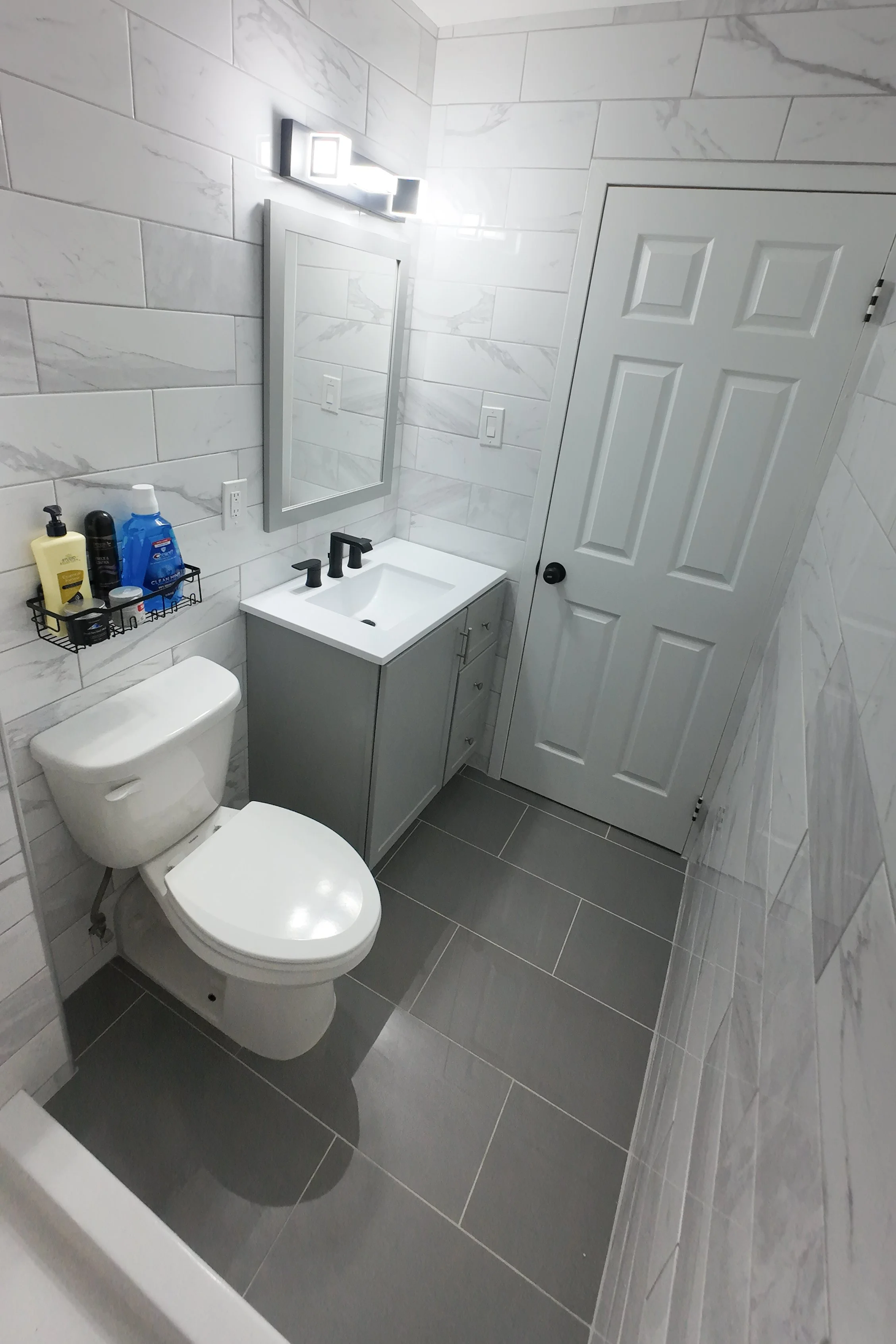 A modern bathroom with gray and white tile walls and gray floor tiles. There is a white toilet and a small gray vanity with a white sink and black fixtures. A mirror and a light fixture are mounted above the sink. A black wire shelf next to the sink 