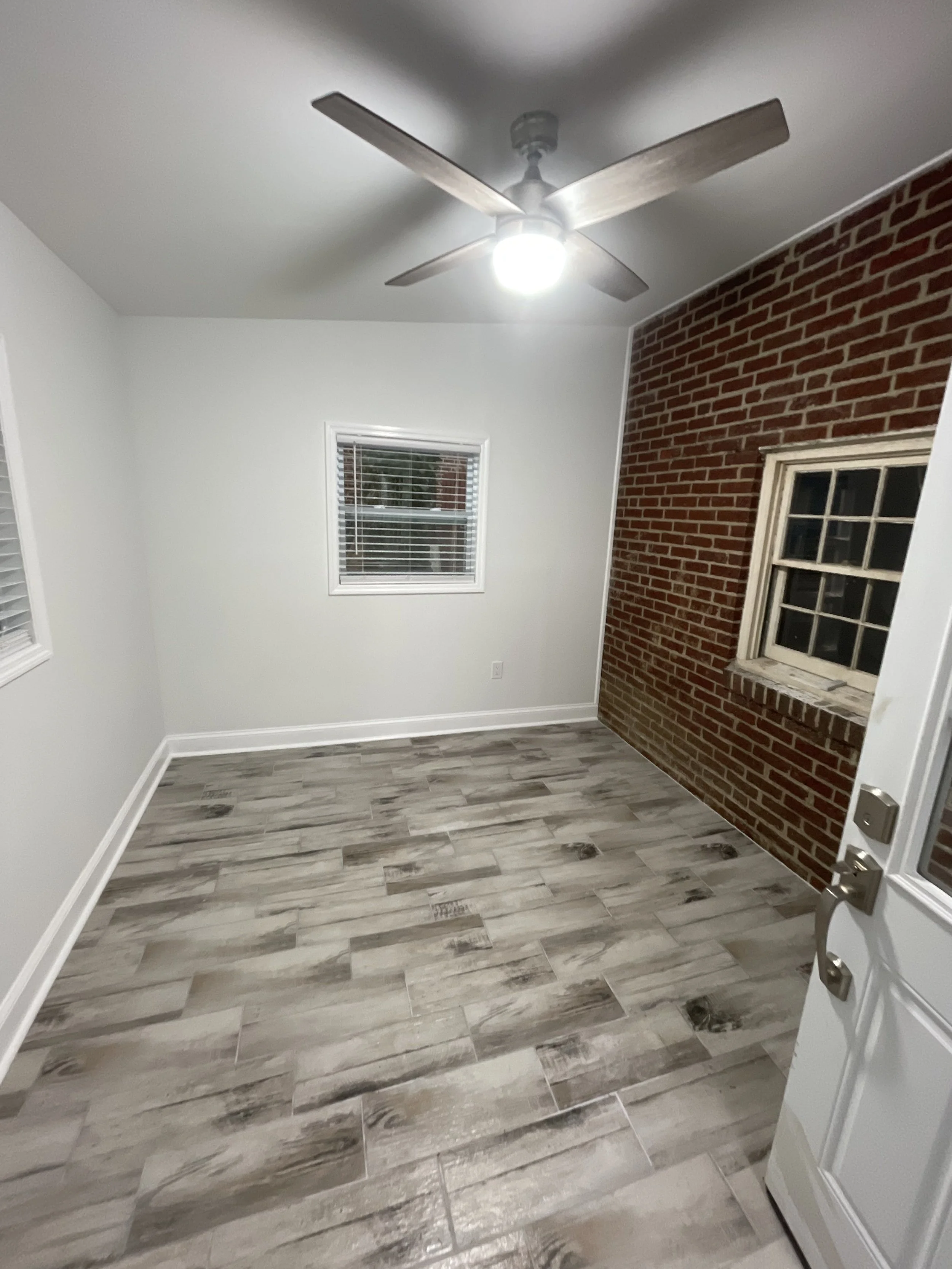 A small room with two windows, white walls, a brick accent wall, and gray wood-look tile flooring, illuminated by a ceiling fan with a light.