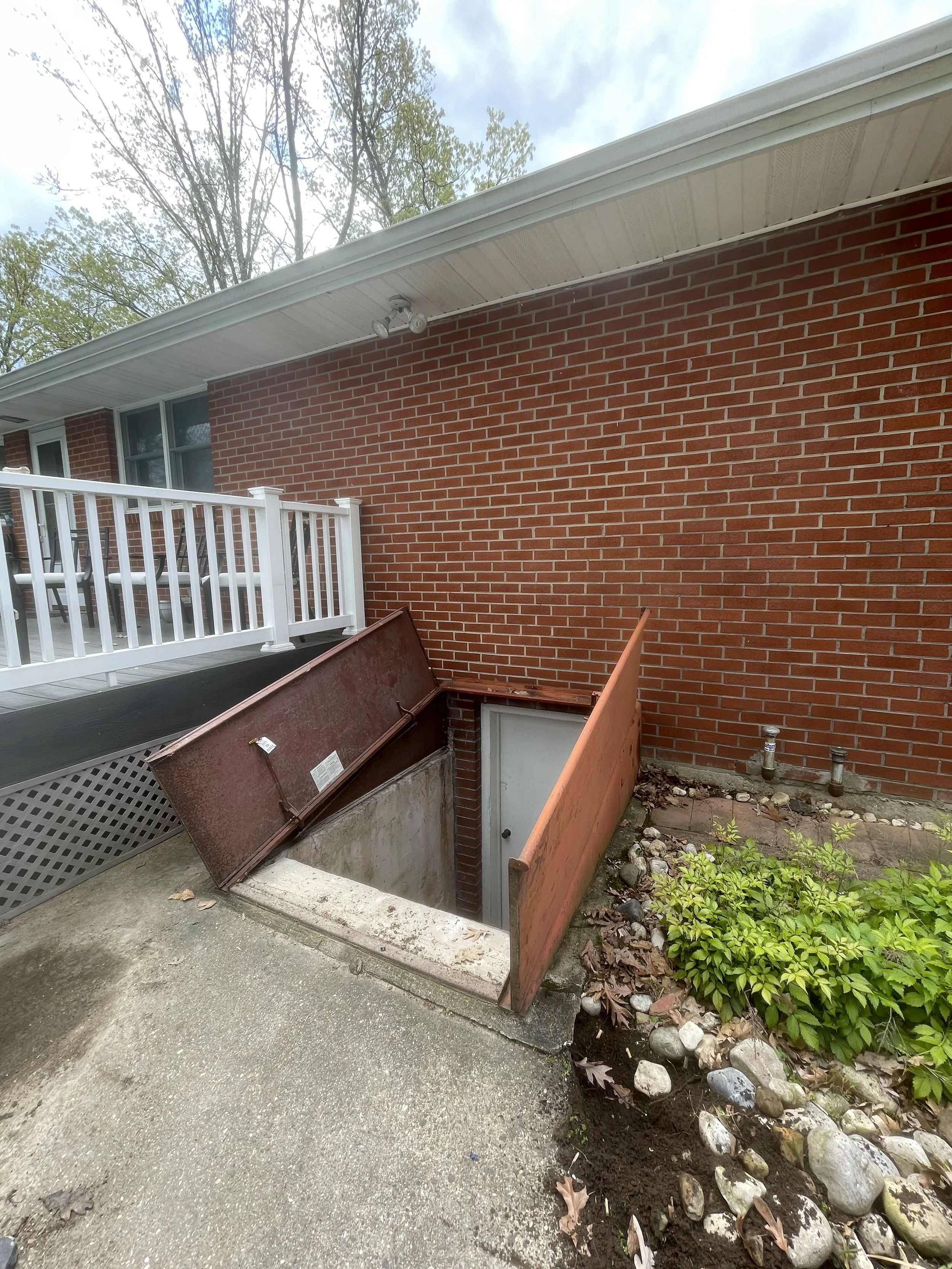 Image of the backyard of a house showing a brick exterior wall, a small door leading to stairs, a potted plant, and some landscaping with rocks and bushes, a white railing, and a vent pipe on the wall.
