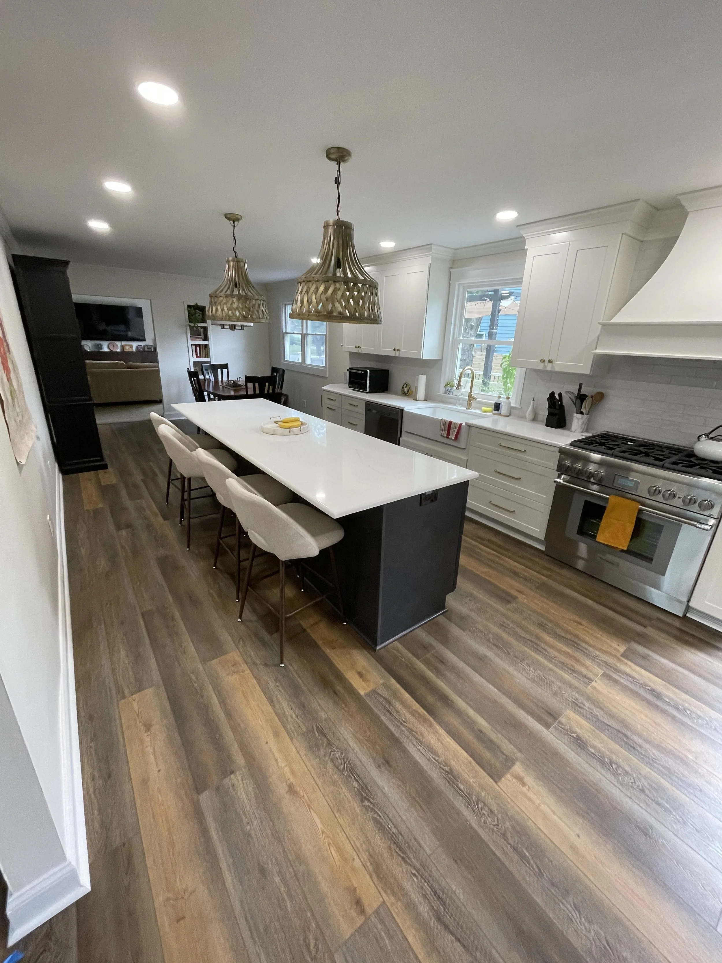 Modern kitchen with white cabinets, stainless steel stove, large white island with beige chairs, wooden flooring, pendant lights, and a window above the sink.