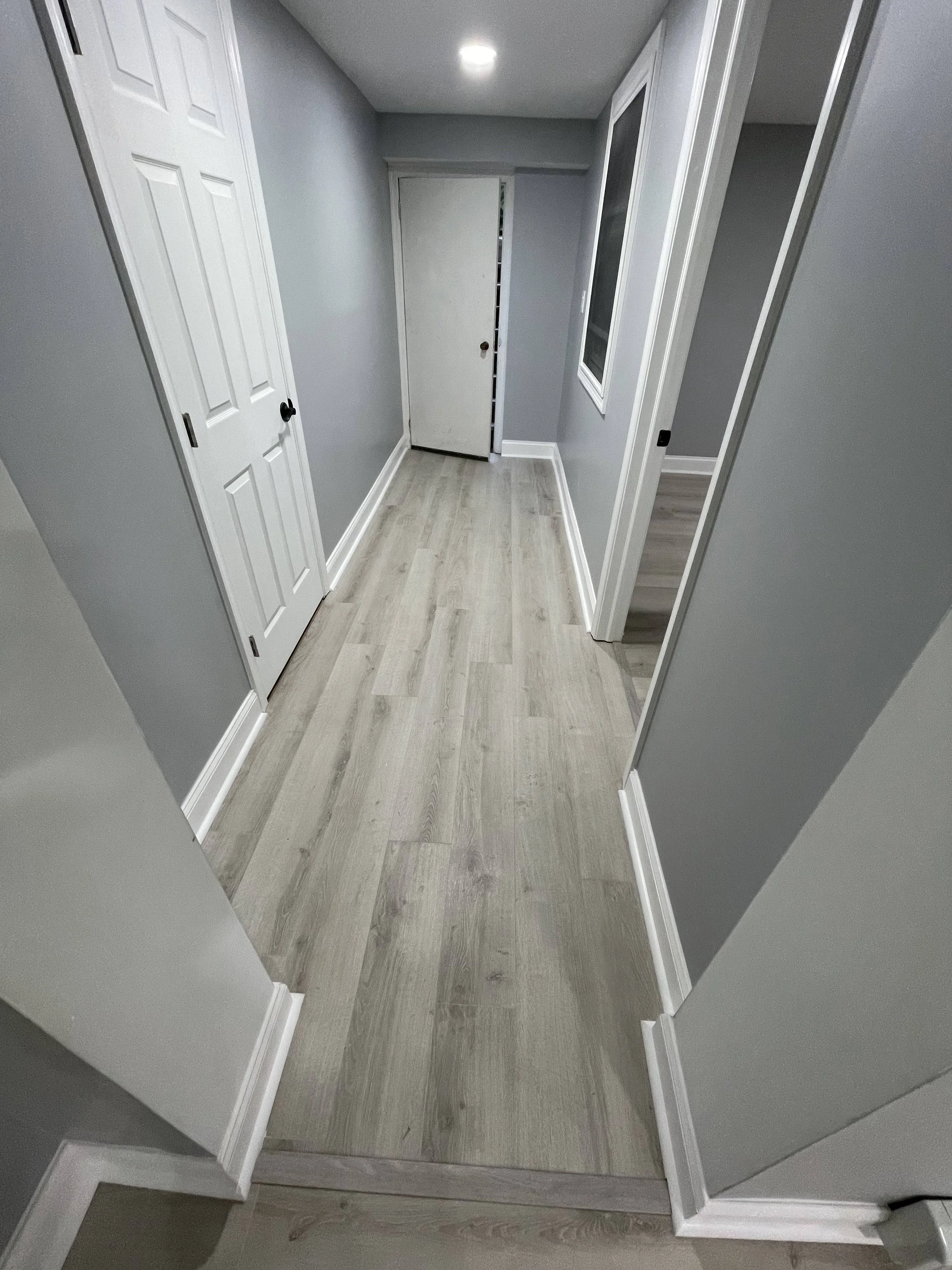 A small hallway with light gray walls, white trim, light wood flooring, and white doors on both sides. The ceiling has recessed lighting.