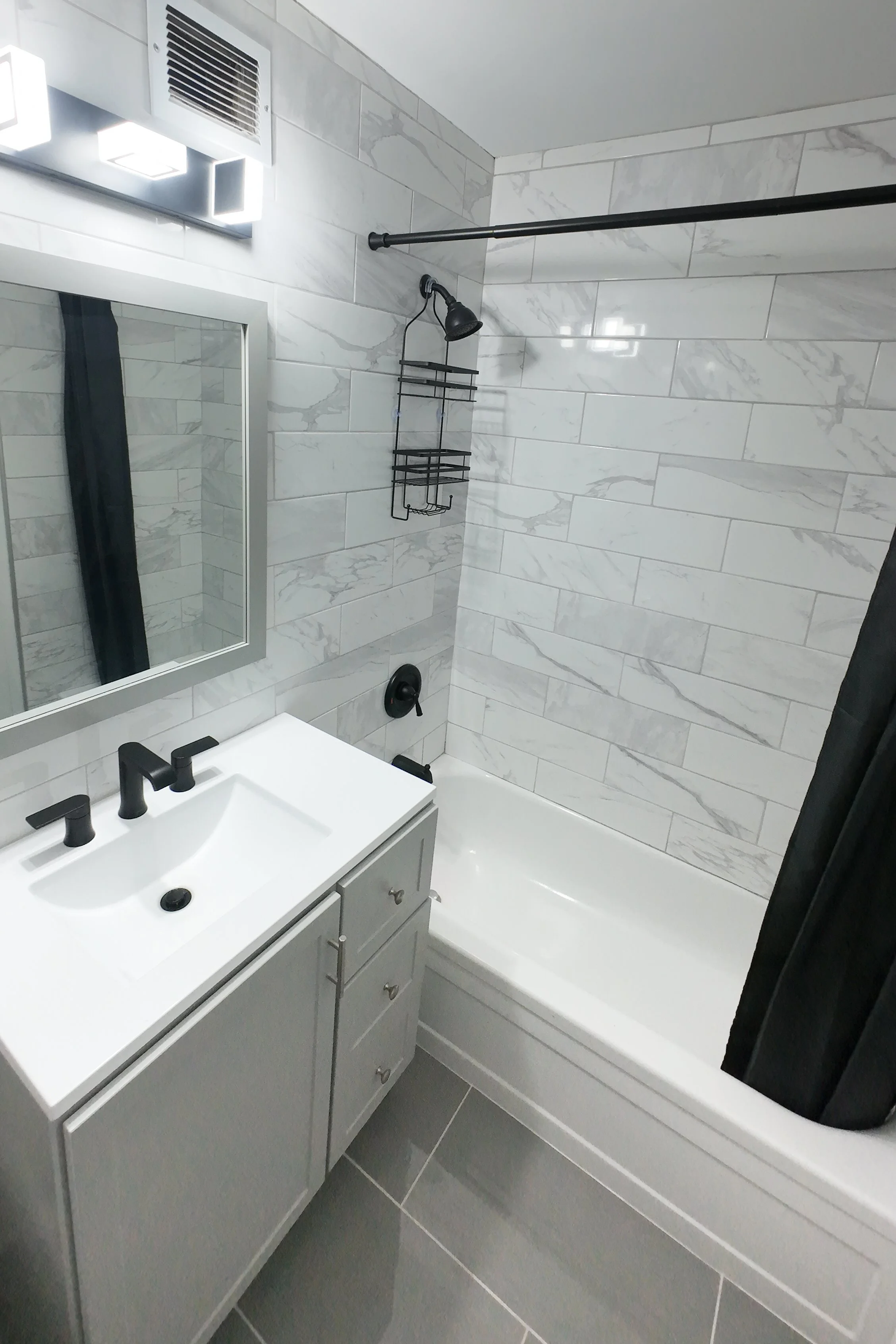 Bathroom with gray vanity, white sink and black fixtures, wall-mounted mirror, gray tiled walls with marble pattern, shower area with black showerhead, shower wall organizer, and gray tiled floor.