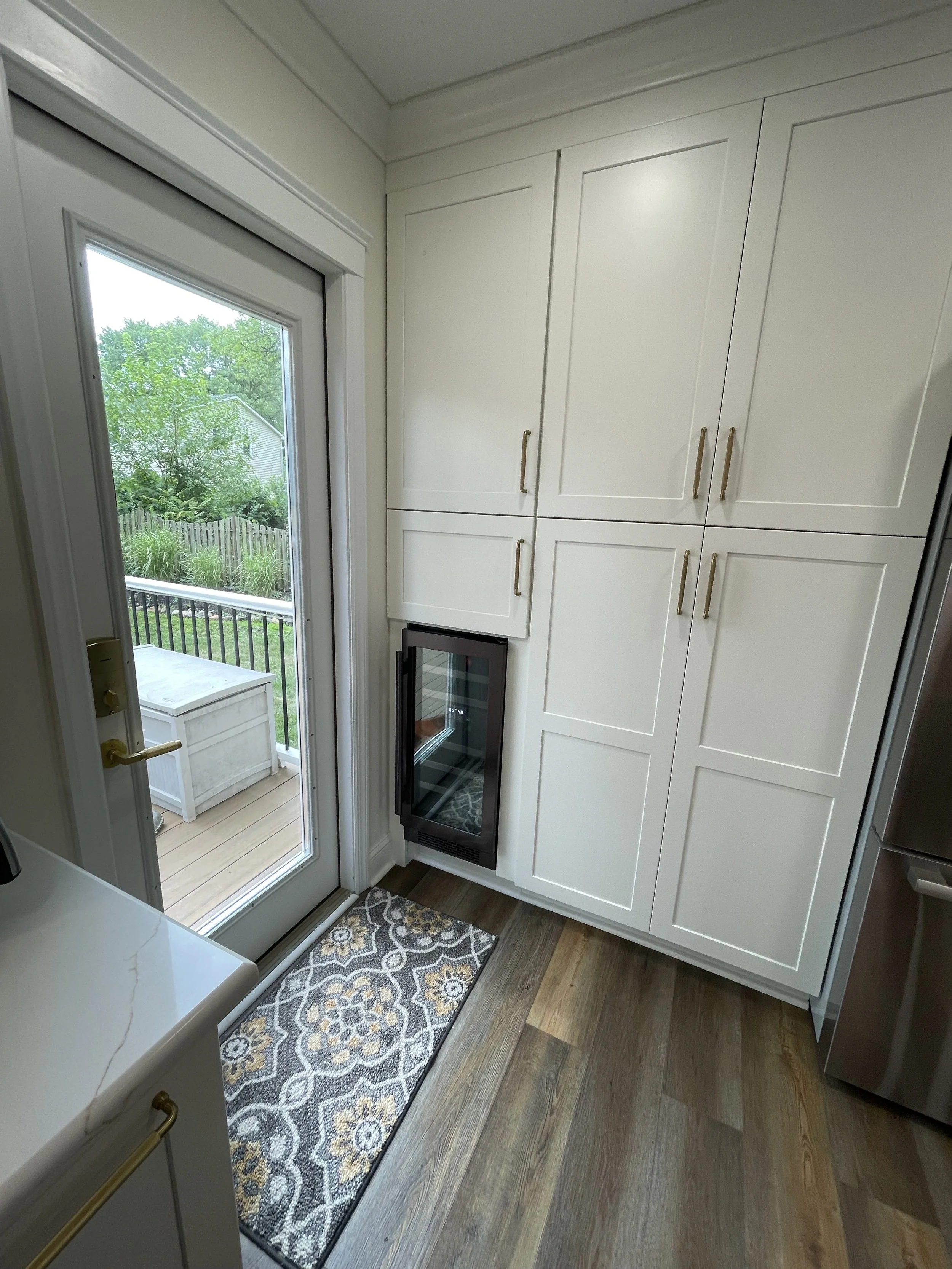 Inside a kitchen with white cabinets, a glass door leading to a balcony with outdoor furniture, a small wine refrigerator, and hardwood flooring with a decorative rug.