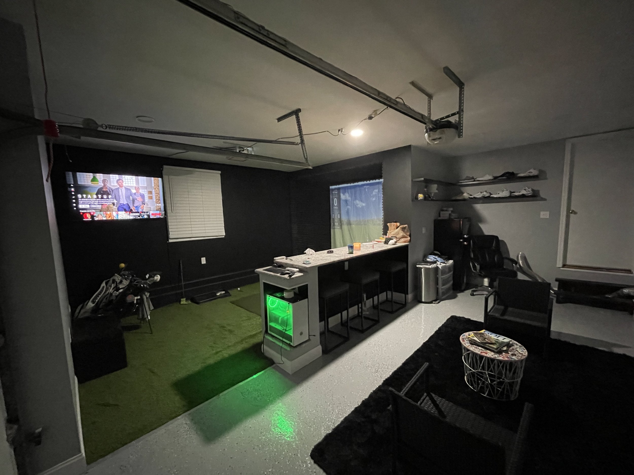 A room with a garage door open, a TV on the wall showing a show, a table with items, and a gaming area with a green-lit gaming computer and golf clubs.