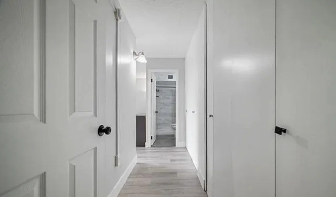Empty hallway with white walls, doors, and wooden flooring leading to a bathroom with marble walls.
