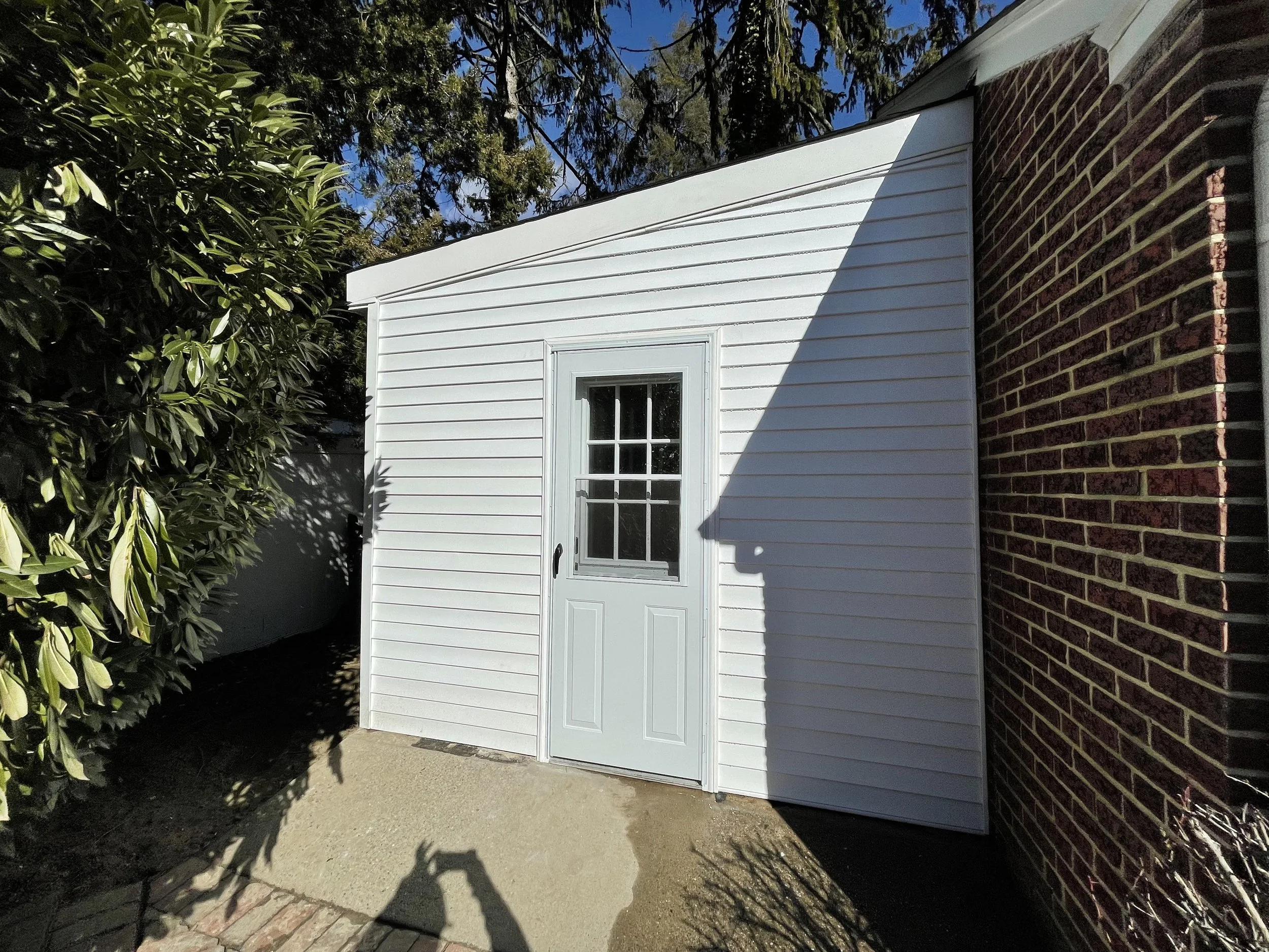 Backyard with white shed, double door, surrounded by trees and bushes, brick wall on one side, concrete patio, shadows cast on ground.