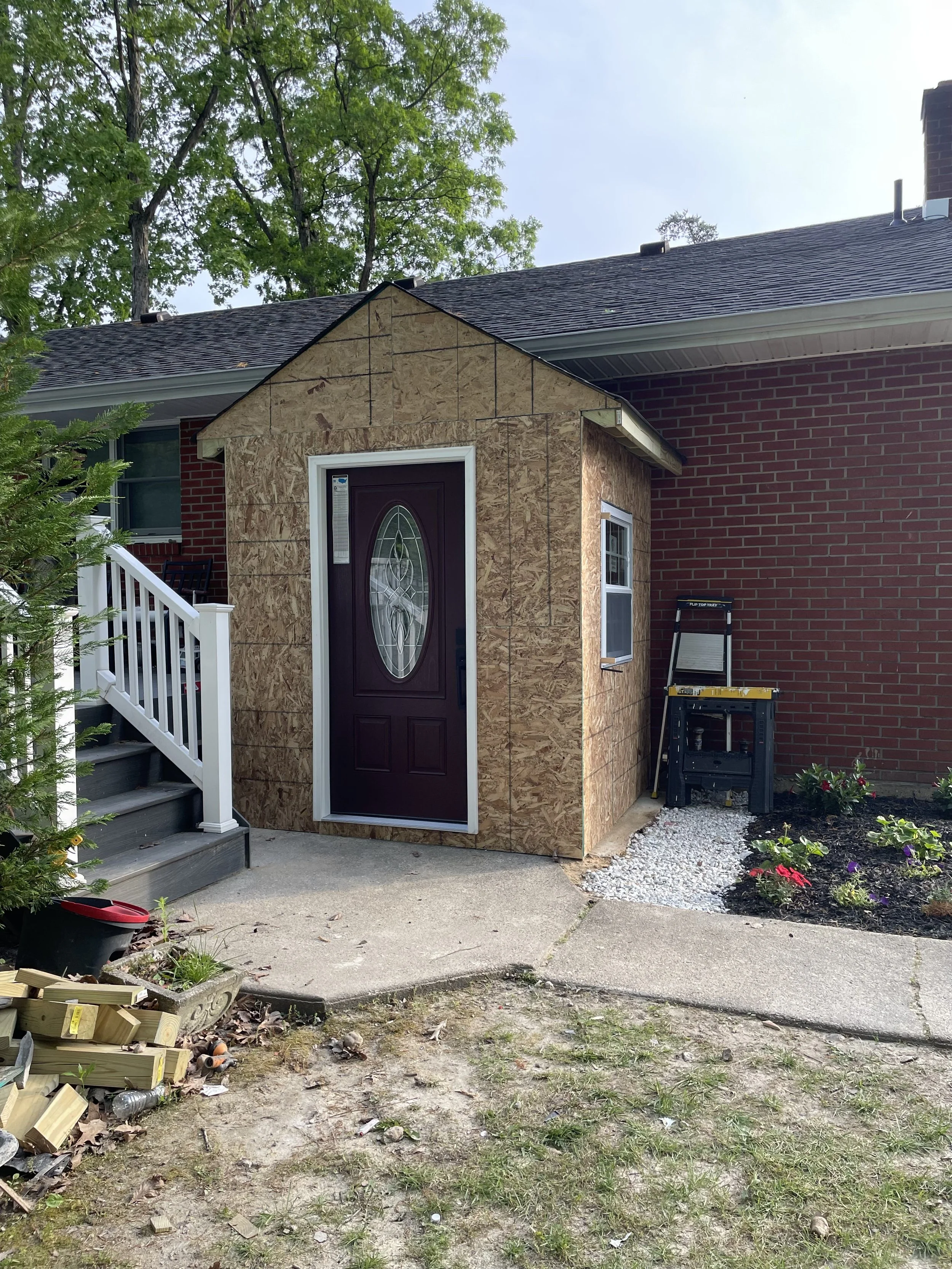 Side view of a house under renovation, with a small structure built in front of the door covered in plywood, a set of stairs leading up to the front door, and construction tools and materials scattered on the ground. There are trees in the background