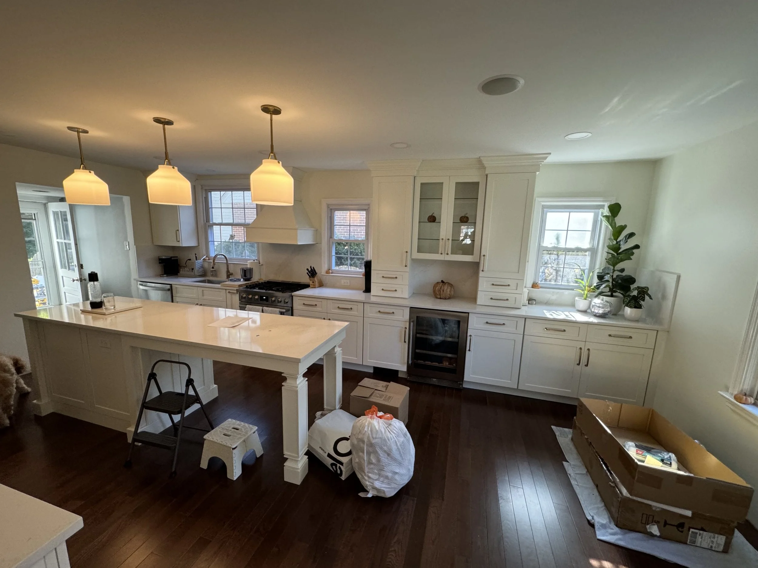 A kitchen with white cabinets, a kitchen island, and three pendant lights hanging from the ceiling. There are two large windows, and various items including boxes and a plant are placed around the room, indicating ongoing organization or setup.