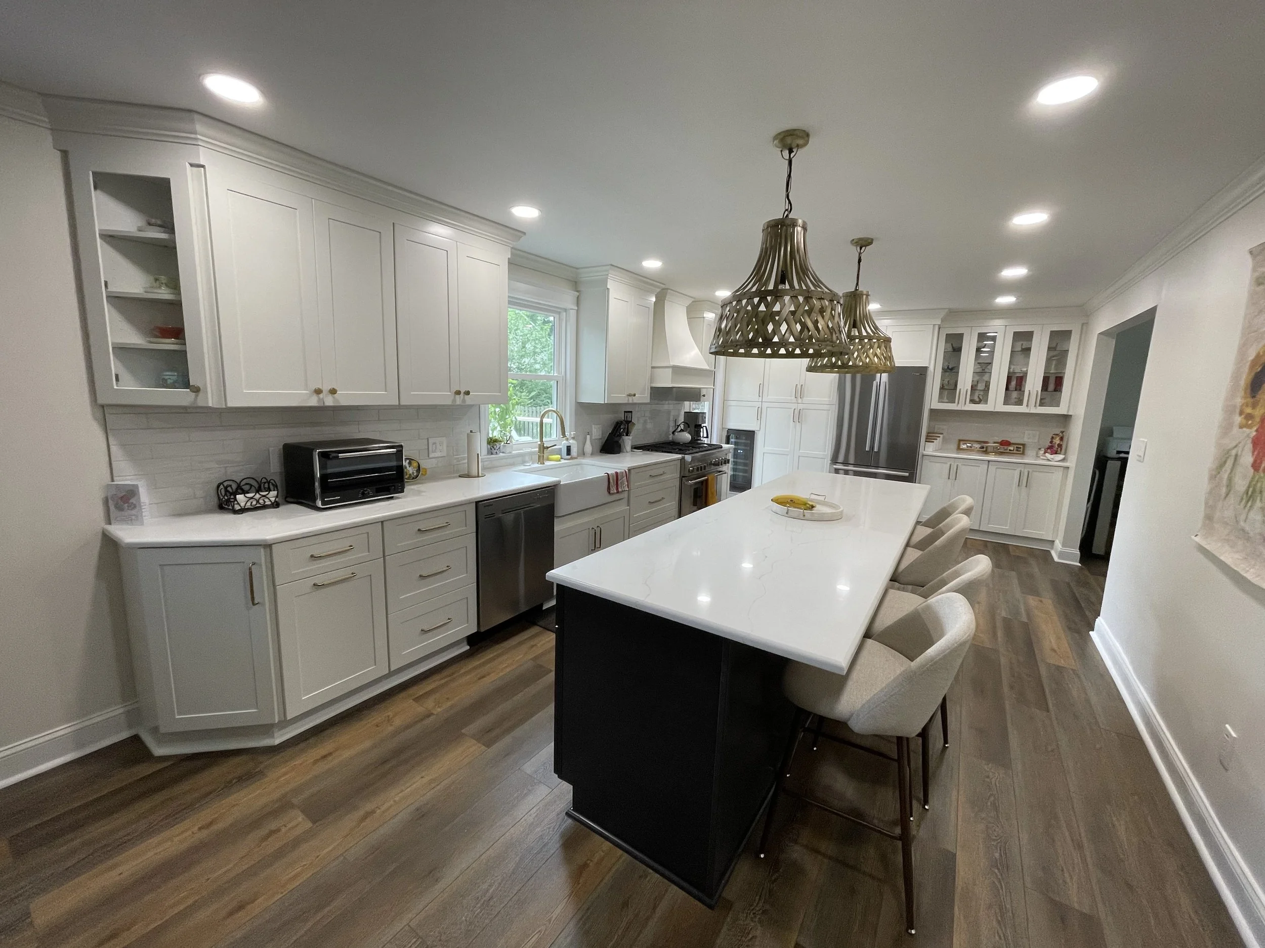 A modern kitchen with white cabinets, a large white center island with black base, pendant lights, stainless steel appliances, hardwood floors, and a window above the sink.