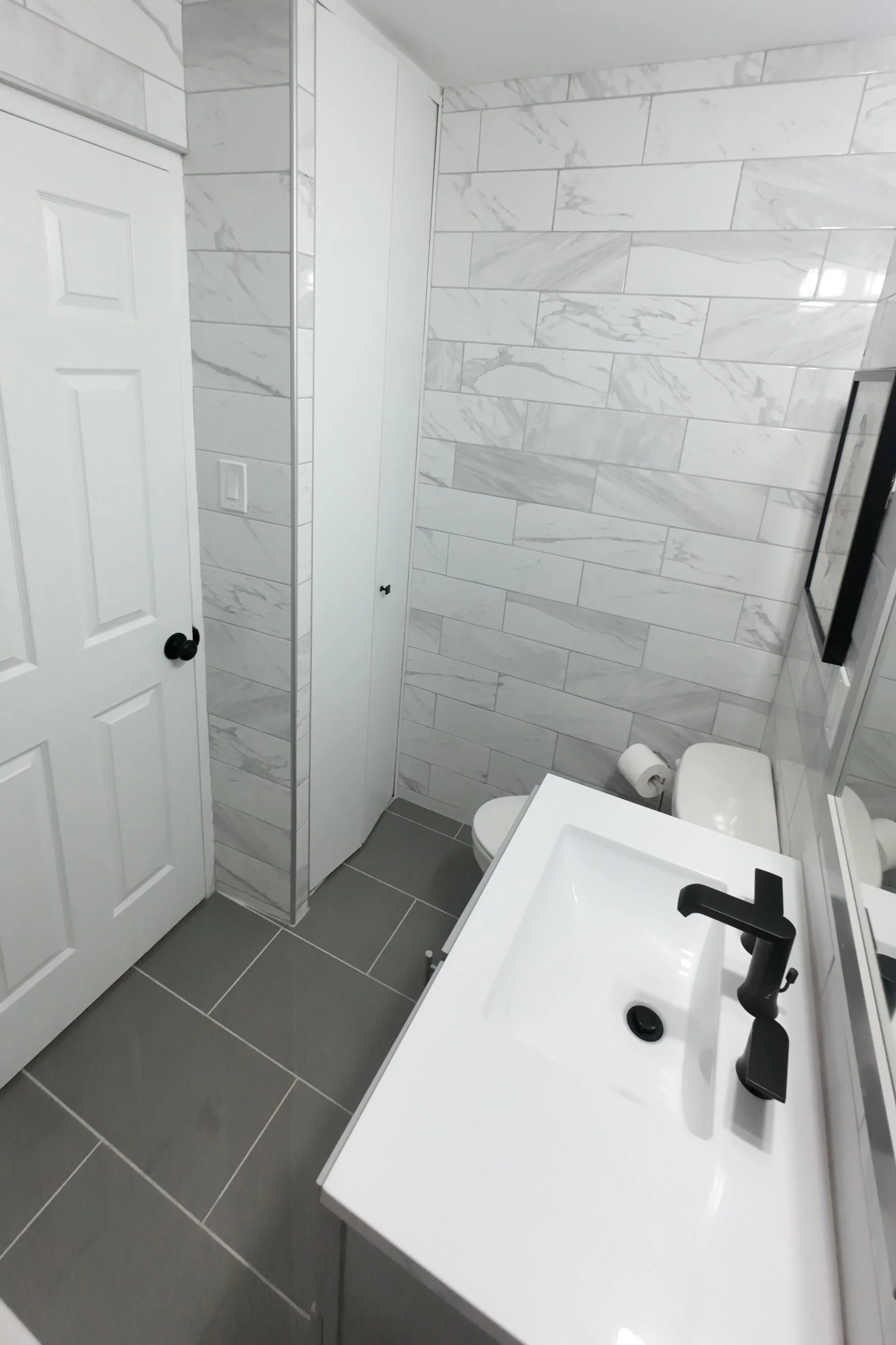 Modern bathroom with gray tiled floor and marble-textured wall tiles, white vanity with black faucet, toilet paper roll, and white cabinetry.