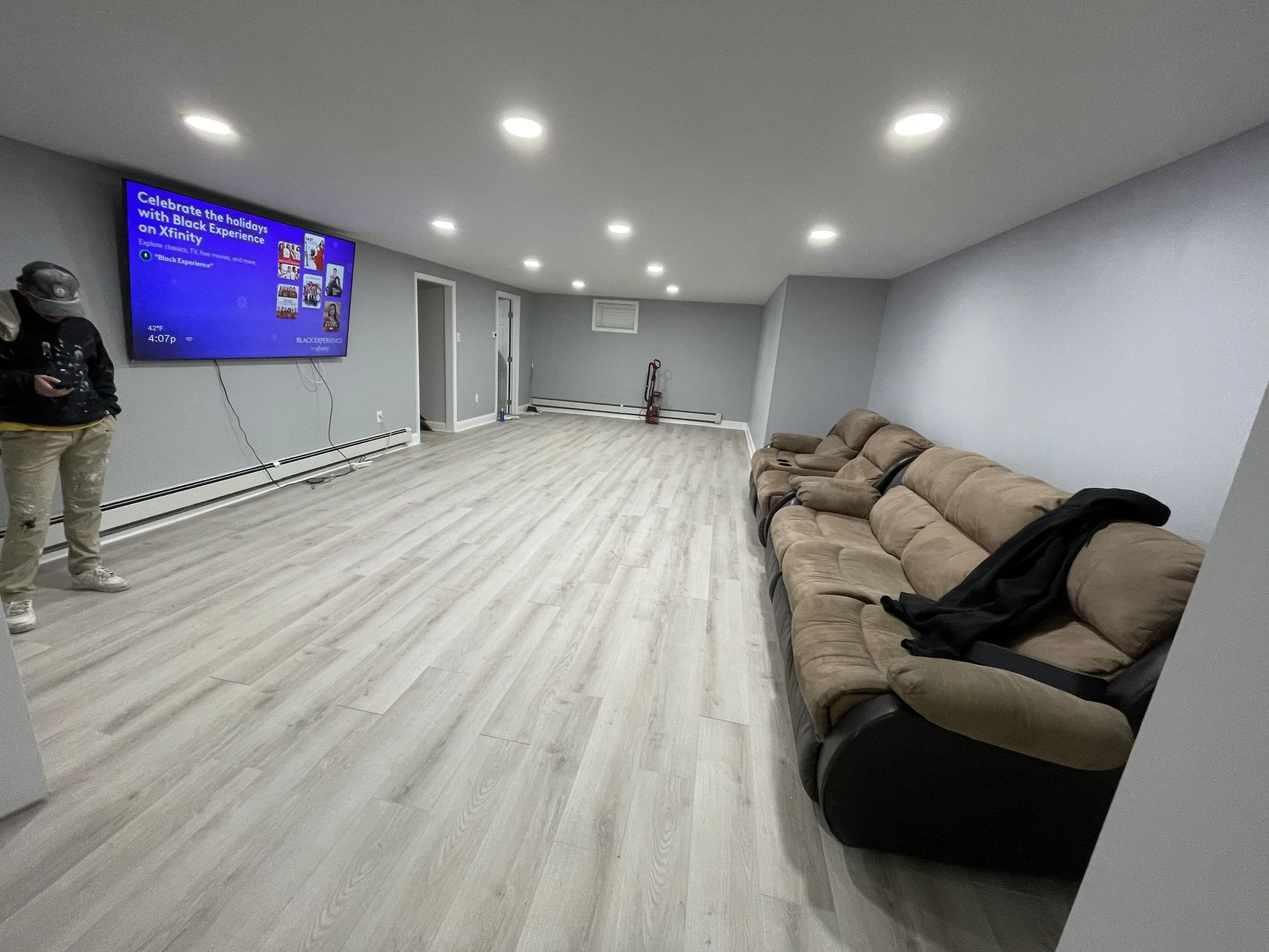 A spacious basement room with light-colored wooden flooring, gray painted walls, and a white ceiling with recessed lighting. There is a flat-screen TV mounted on the wall on the left, displaying a promotional slide for Black Experience on Xfinity. A 
