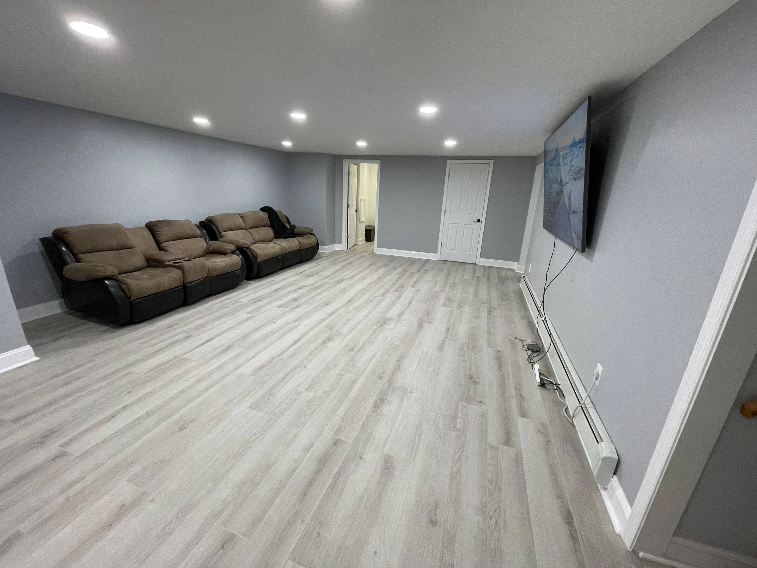 Living room with grey walls, recessed ceiling lights, a flat-screen TV mounted on the wall, tan recliner sofas, light wood flooring, and closed doors.