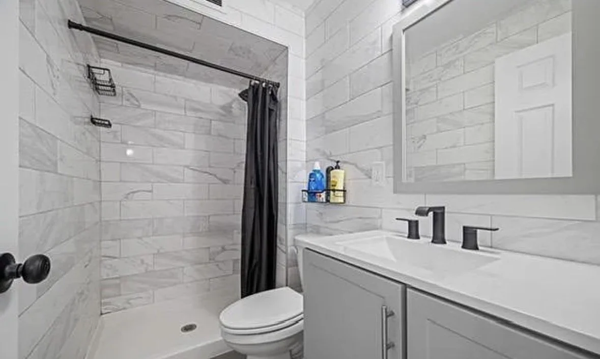 Bathroom with marble walls, a shower area with a black shower curtain, a toilet, and a white vanity with a black faucet and mirror.
