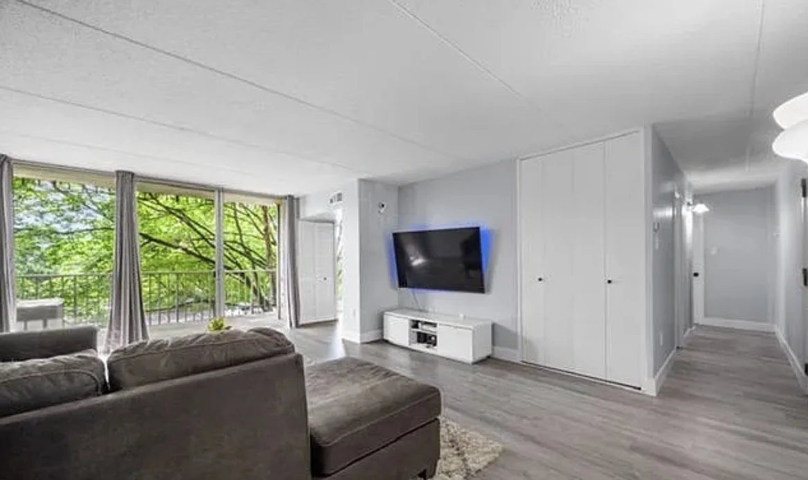 Living room with gray sofa, wood flooring, large windows with trees outside, TV on white console, hallway with white doors.