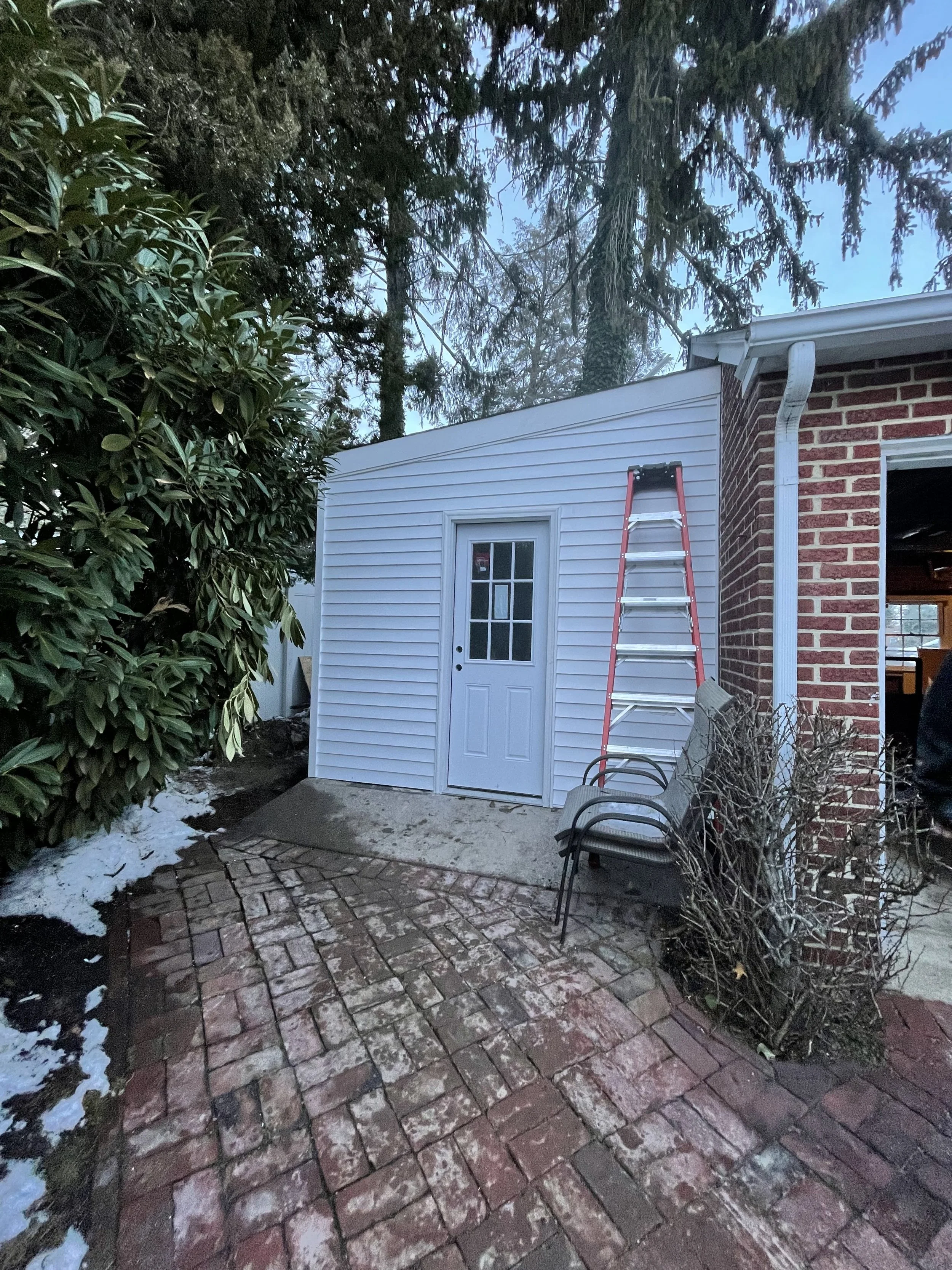 Backyard patio with a brick paver floor, a white shed, a red step ladder, some chairs, and nearby trees and bushes with patches of snow on the ground.