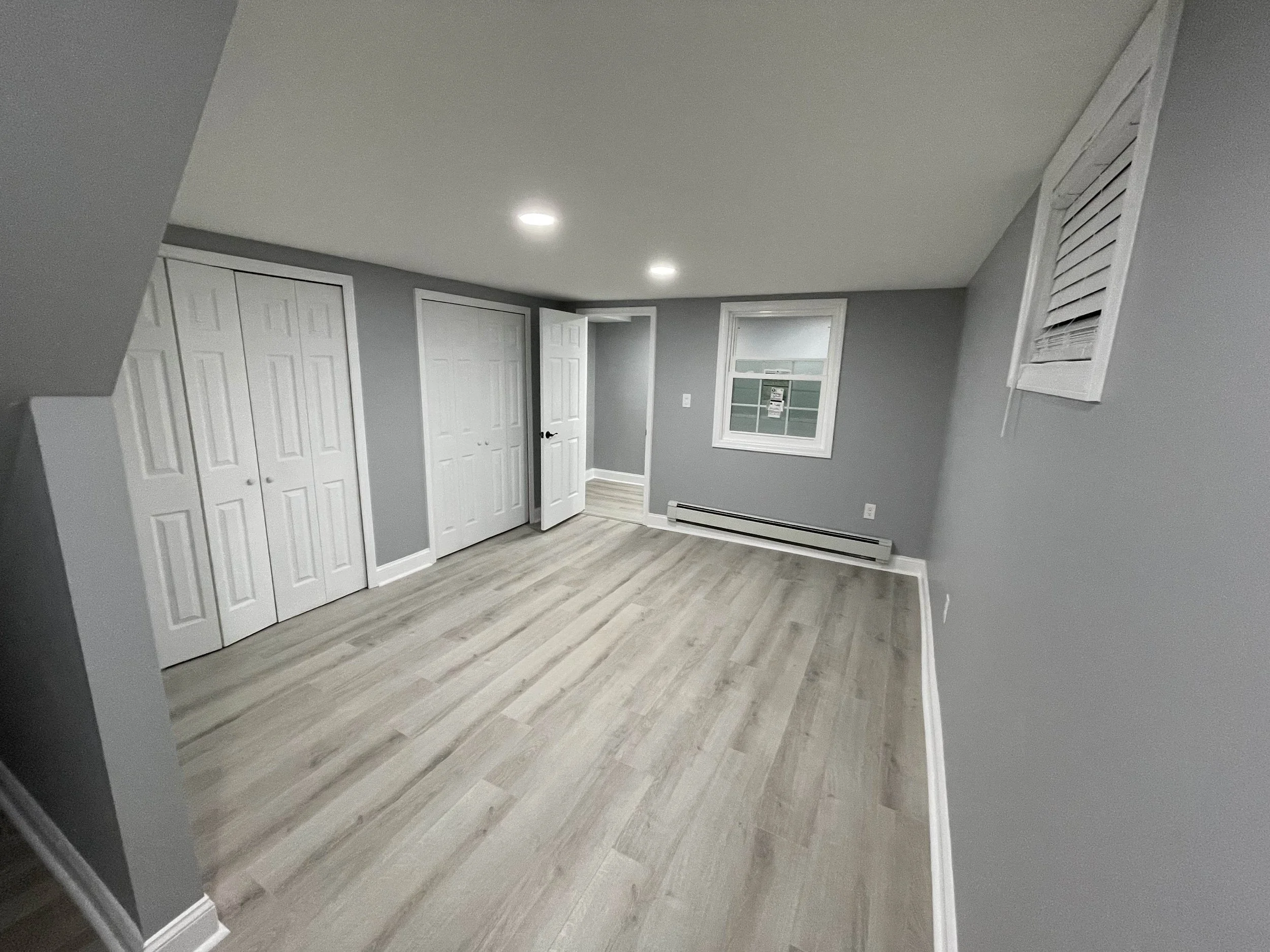 Room with light-colored wood flooring, gray walls, two ceiling lights, a window, white closet doors, and an air vent on the wall.