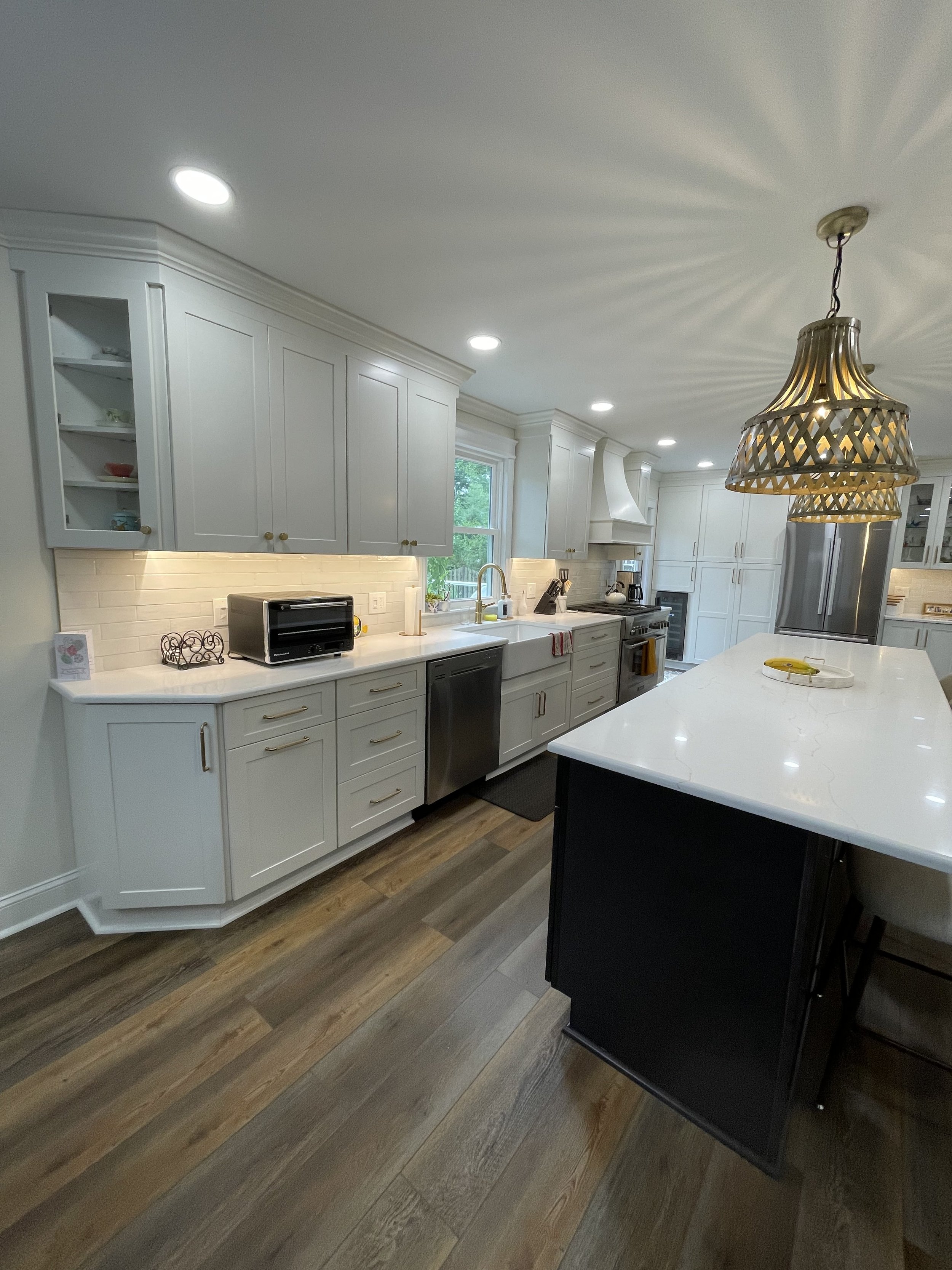 Modern kitchen with white cabinets, stainless steel appliances, and a large white island counter. Recessed lighting and a hanging pendant light fixture above the island. Wood flooring and a window over the sink.