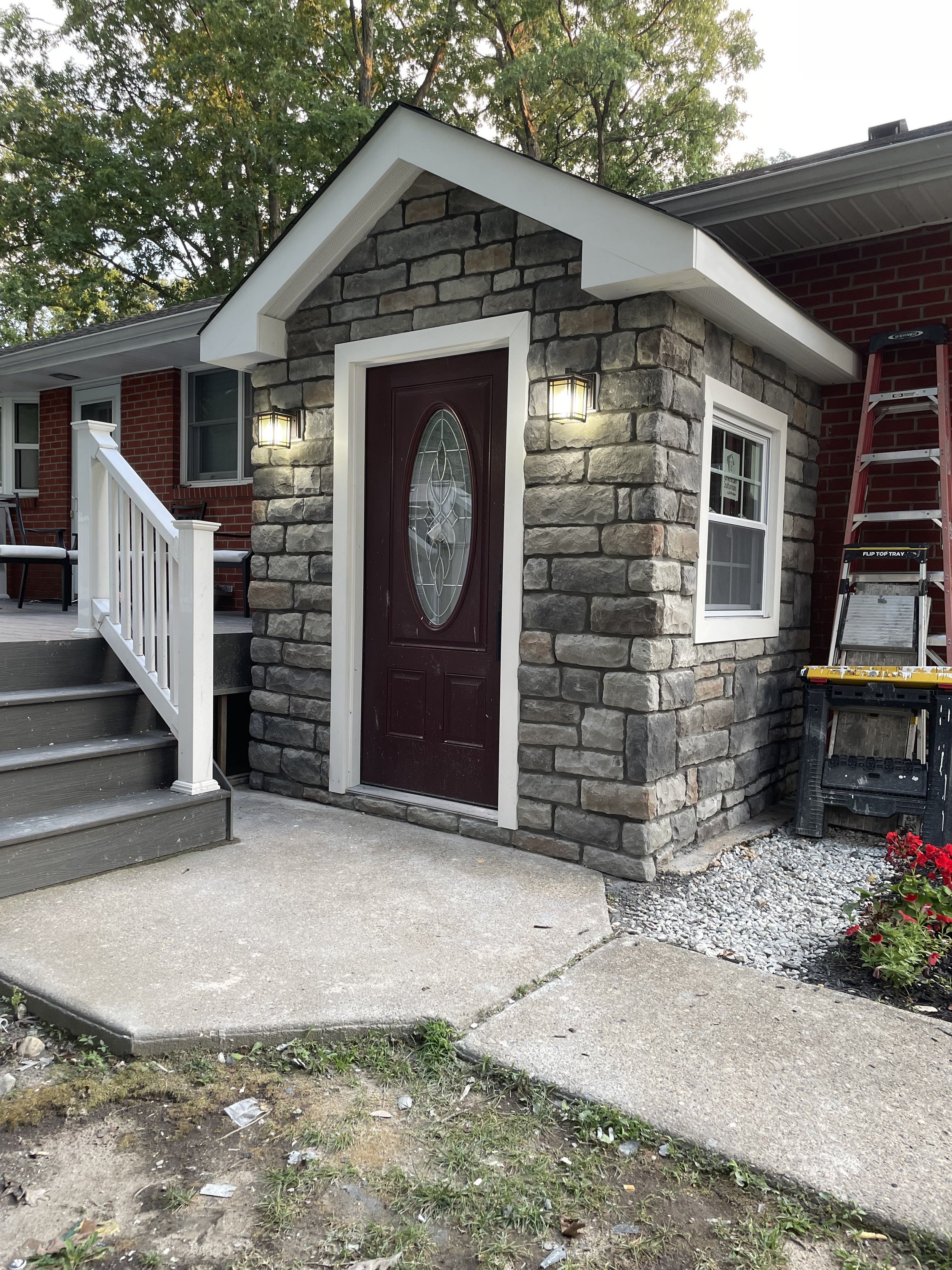 Front entrance of a house with a stone facade, a dark wooden door with glass design, two wall-mounted lamps, a small porch with stairs, and a ladder leaning against the house. There are trees and a lawn in the background, and some construction or lan