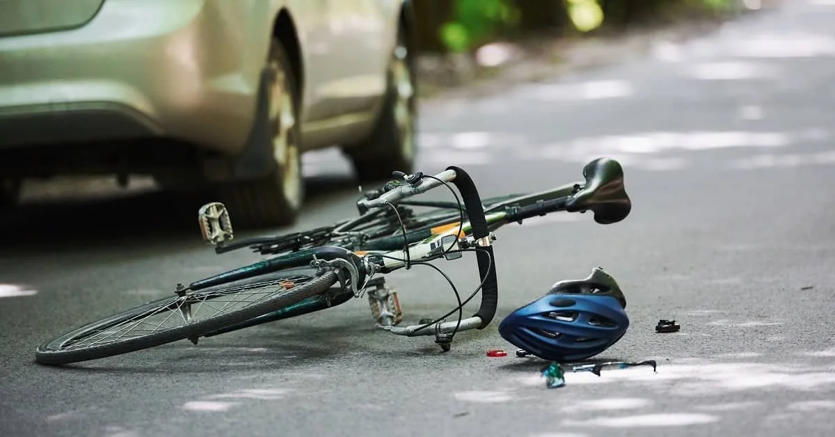 A bicycle lying on its side on the road next to a car, with a blue helmet and other small items nearby, indicating a possible accident.