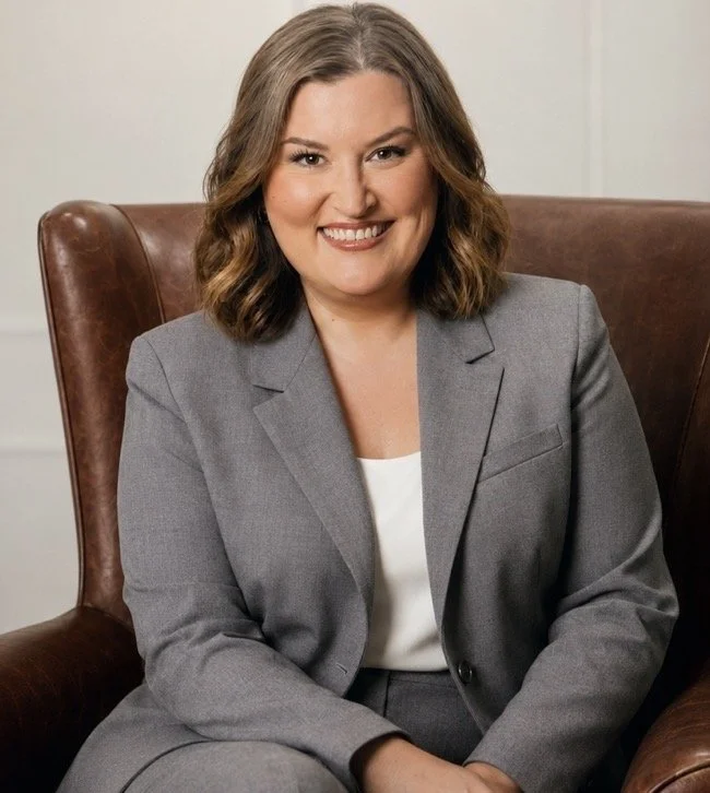 A woman with shoulder-length wavy brown hair and a warm smile, wearing a gray blazer over a white top, sitting in a brown leather armchair.